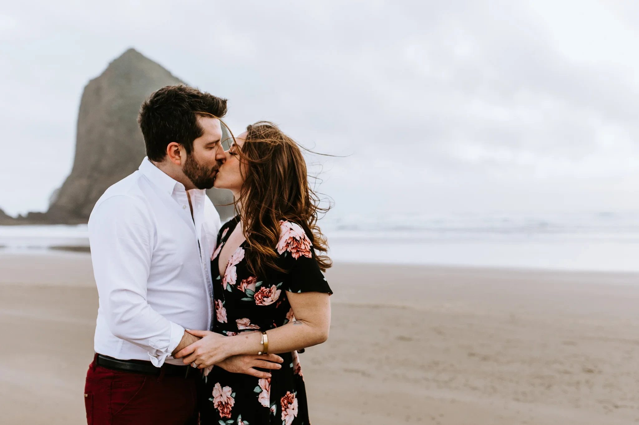 Two adults kiss in front of a distant sea stack