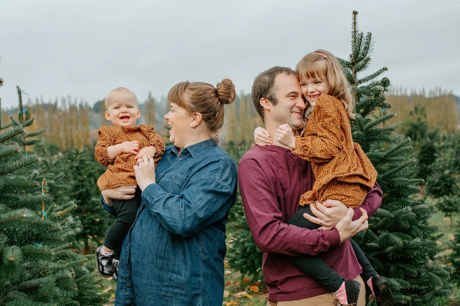 Two adults hold two smiling children among rows of evergreen trees