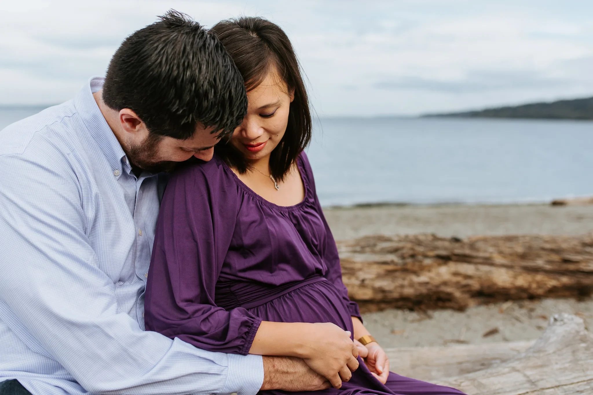 Two adults sit close together at the shoreline, one wearing a purple dress