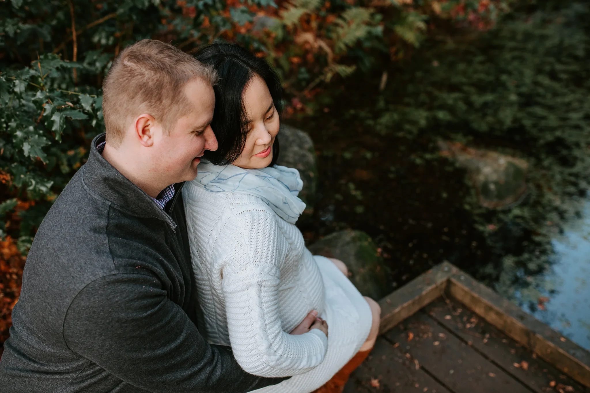 Two adults lean together on a wooden walkway beneath evergreen trees