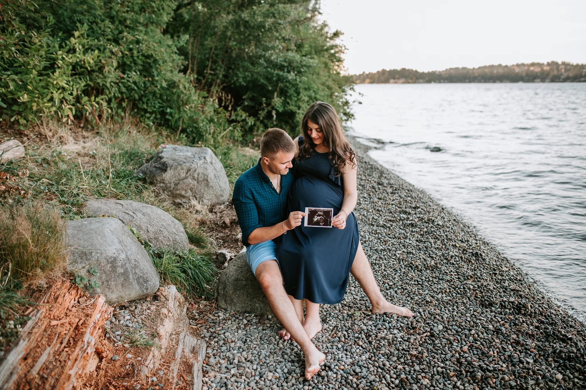 Two adults sit on shoreline rocks beside calm water