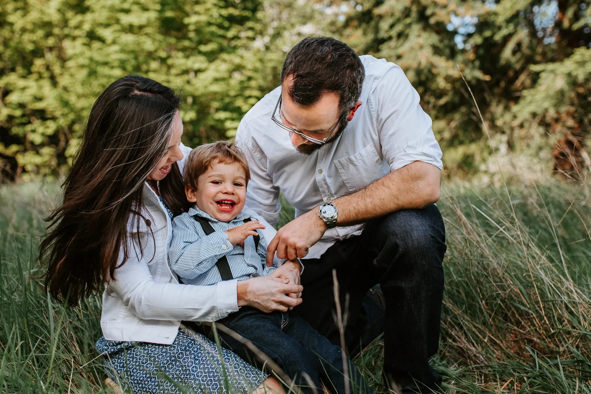 Two adults laugh with a toddler while sitting in tall grass