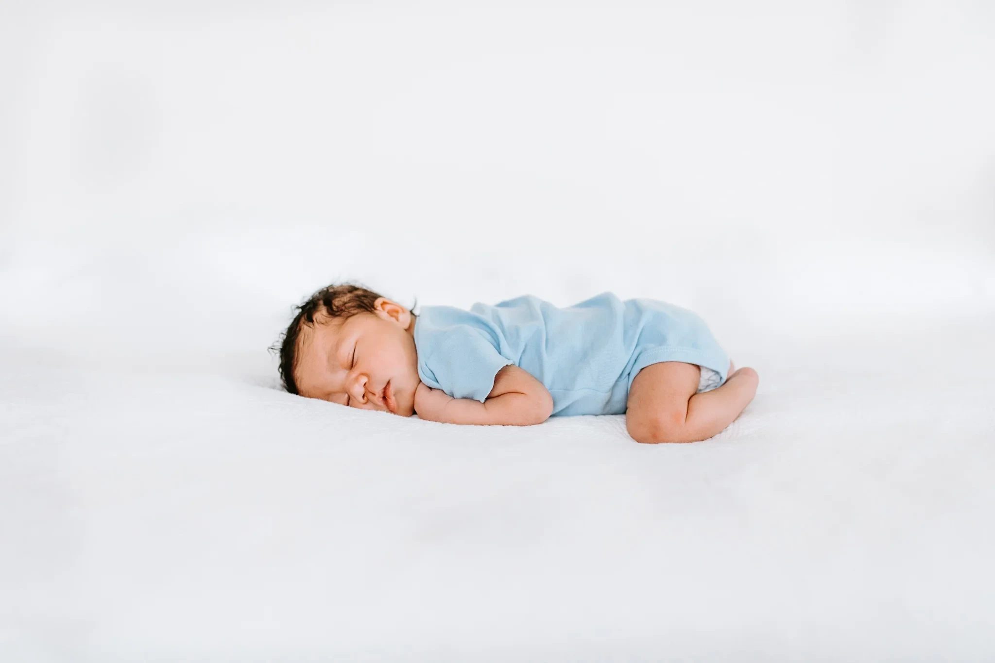 Sleeping baby in a pale blue outfit lies on white bedding