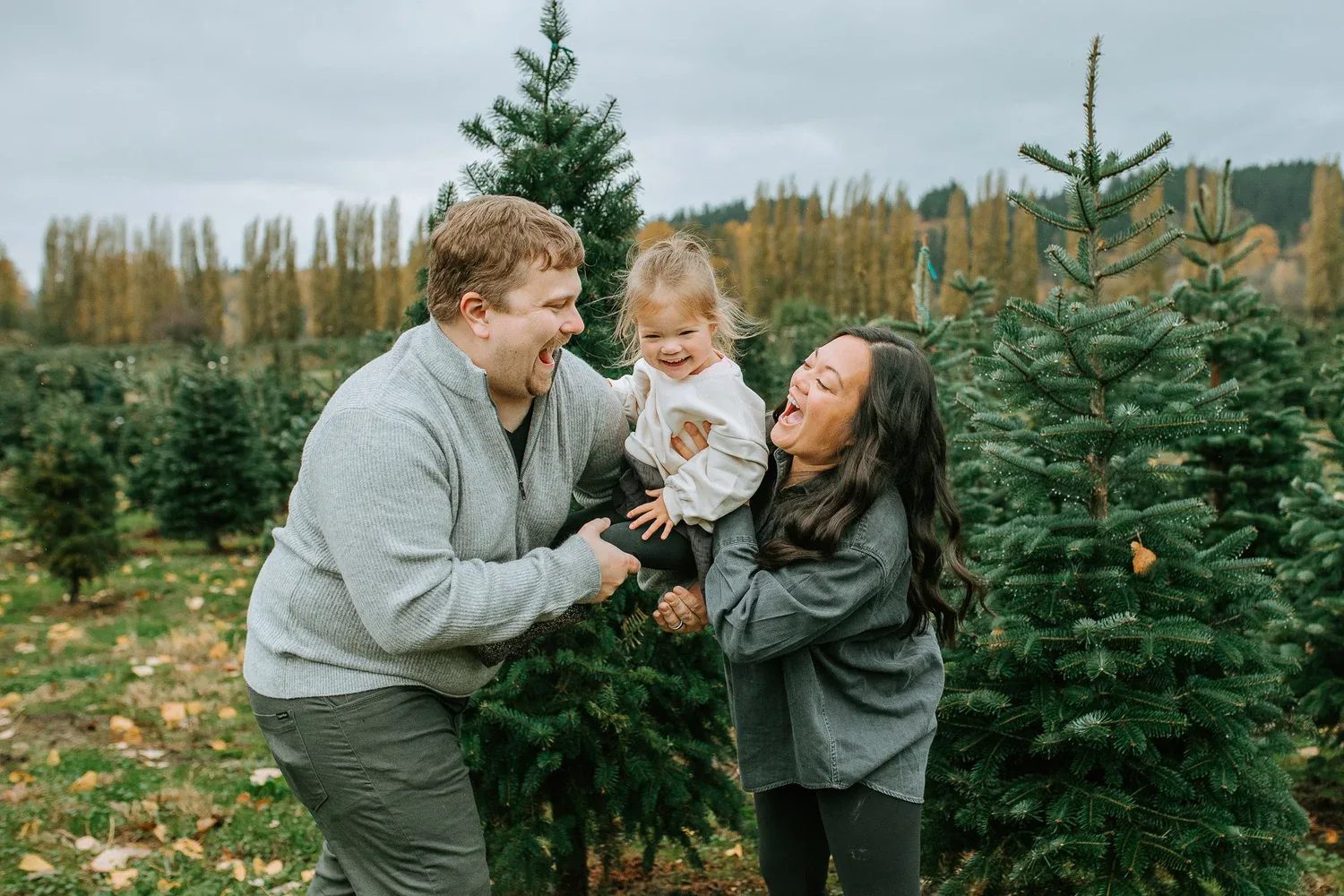 Two adults kiss a toddler’s cheeks at a tree farm