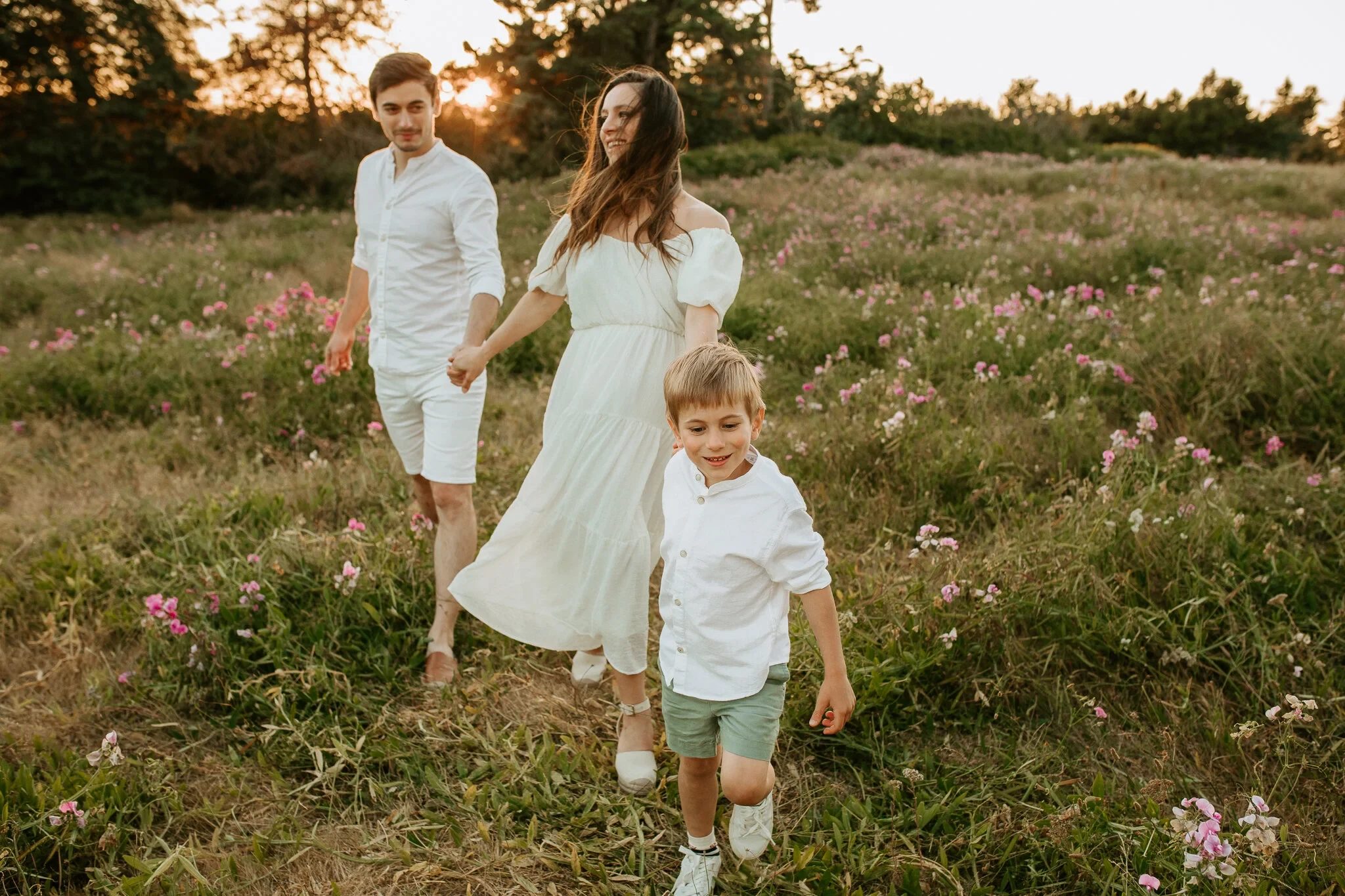 Two adults and a child in light clothing walk through a sunlit meadow