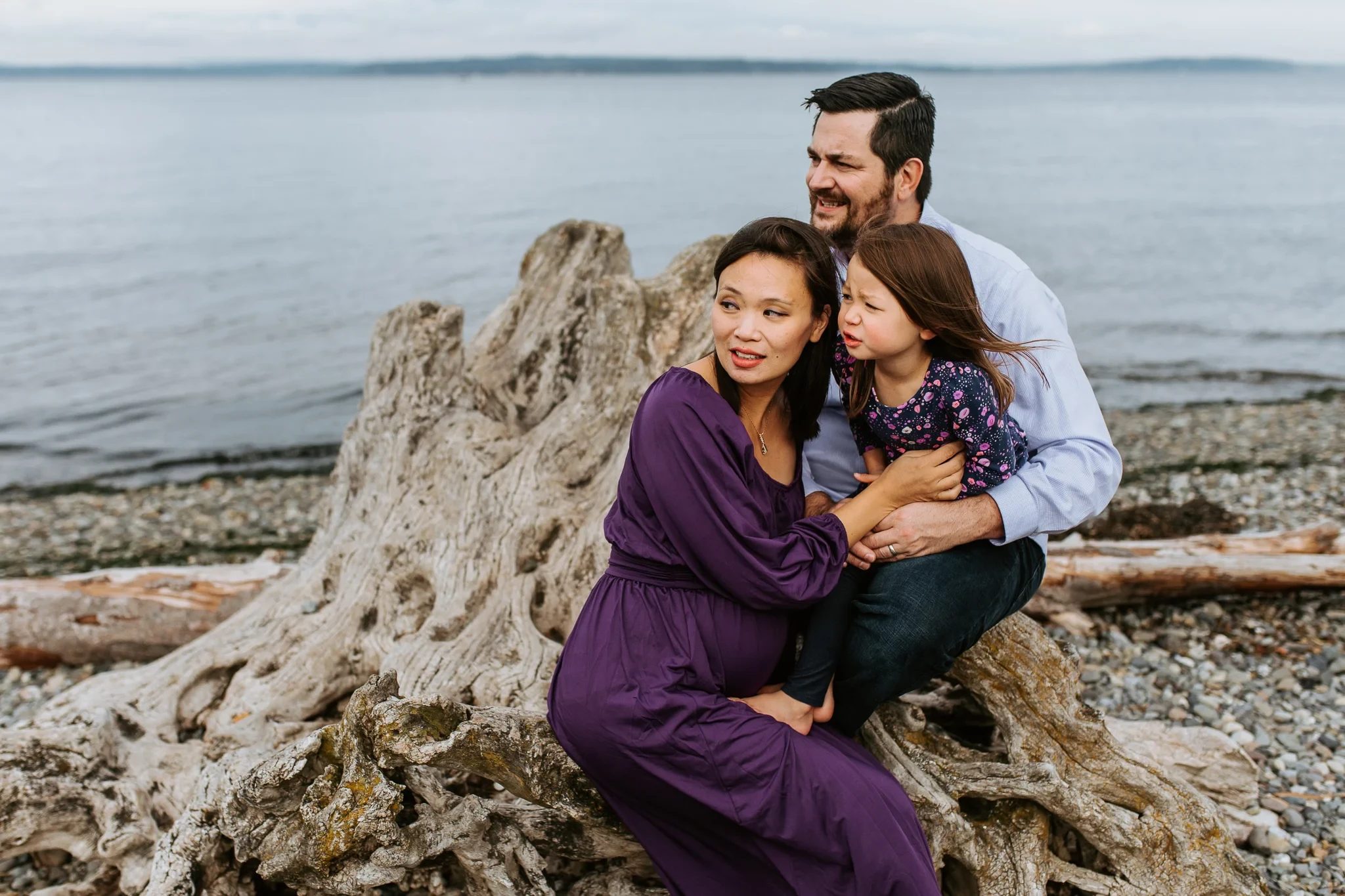 Two adults and a child sit together beside driftwood on the beach
