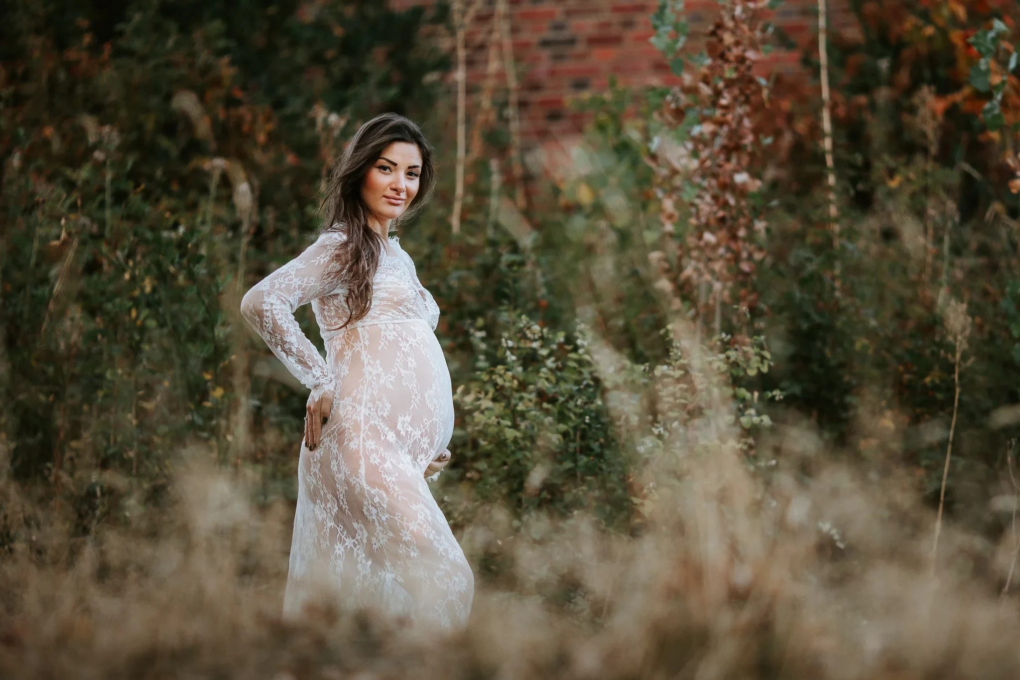 Woman in a pale lace dress stands among tall grass and leafy shrubs