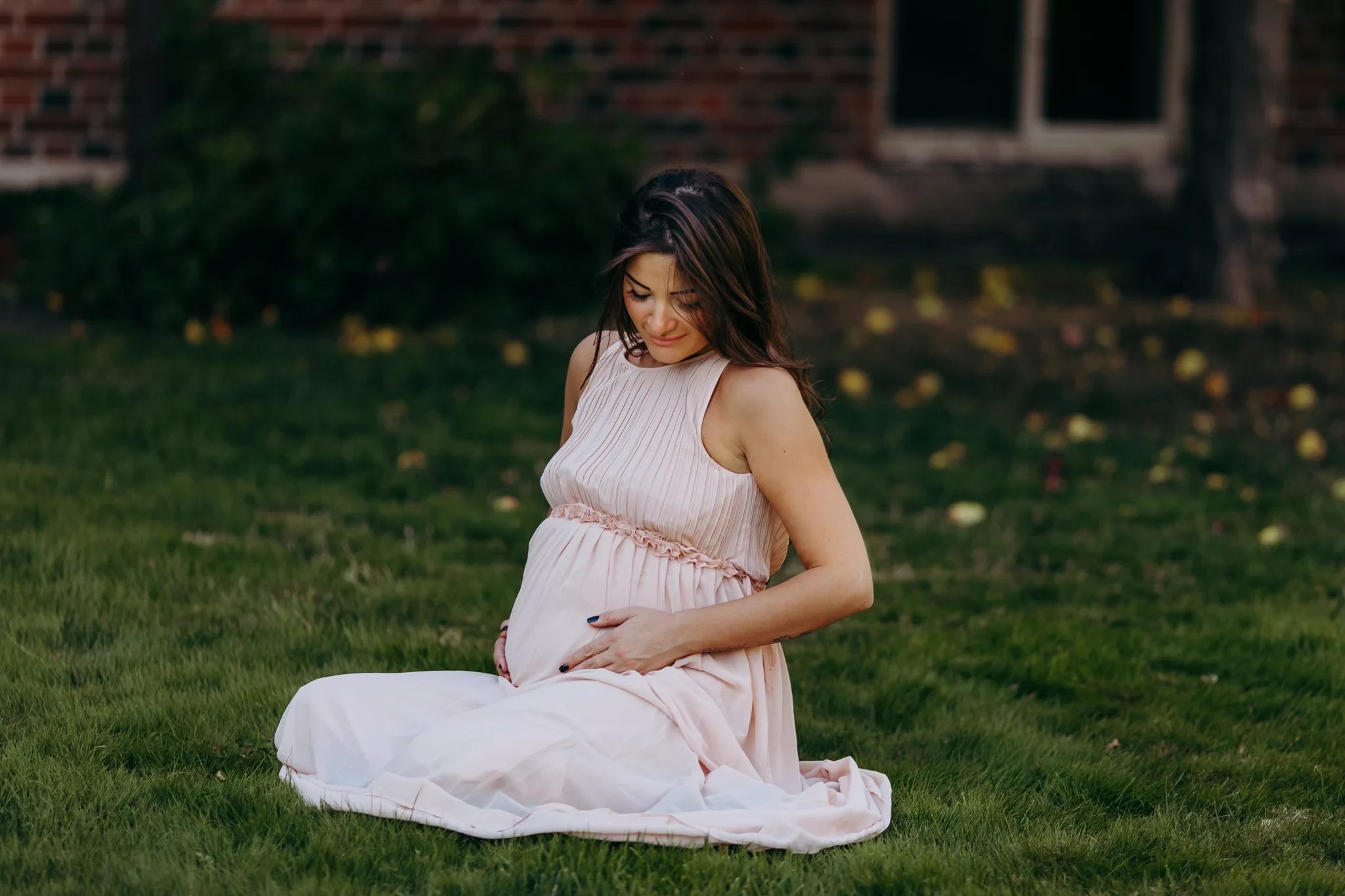 Woman in a pink dress sits on a lawn in front of a brick building