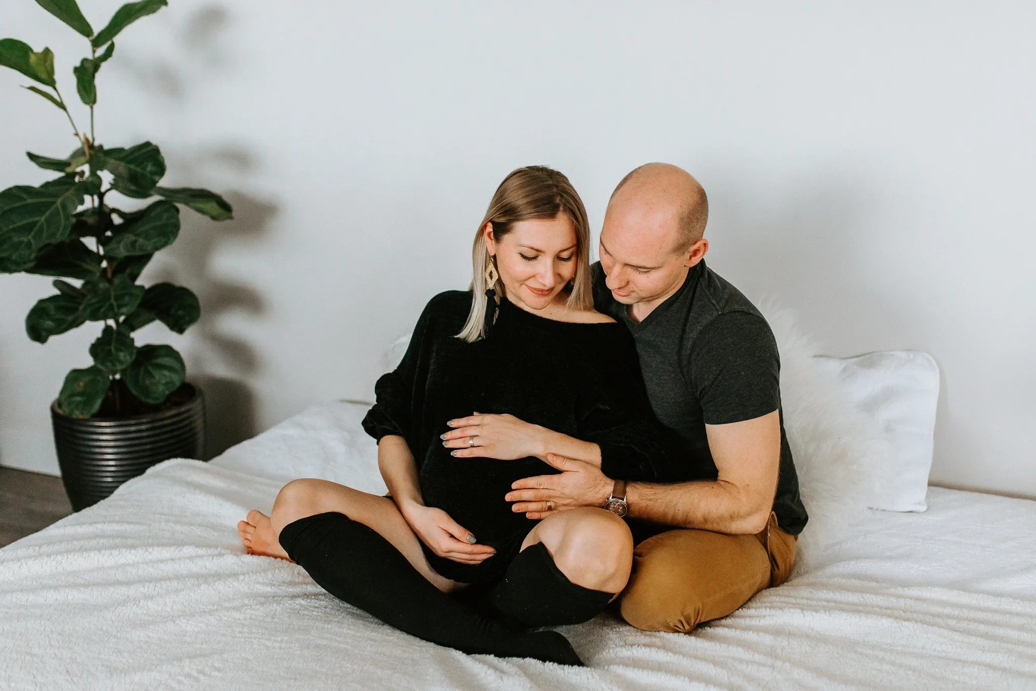 Two adults sit cross-legged together on a white bed beside a leafy plant