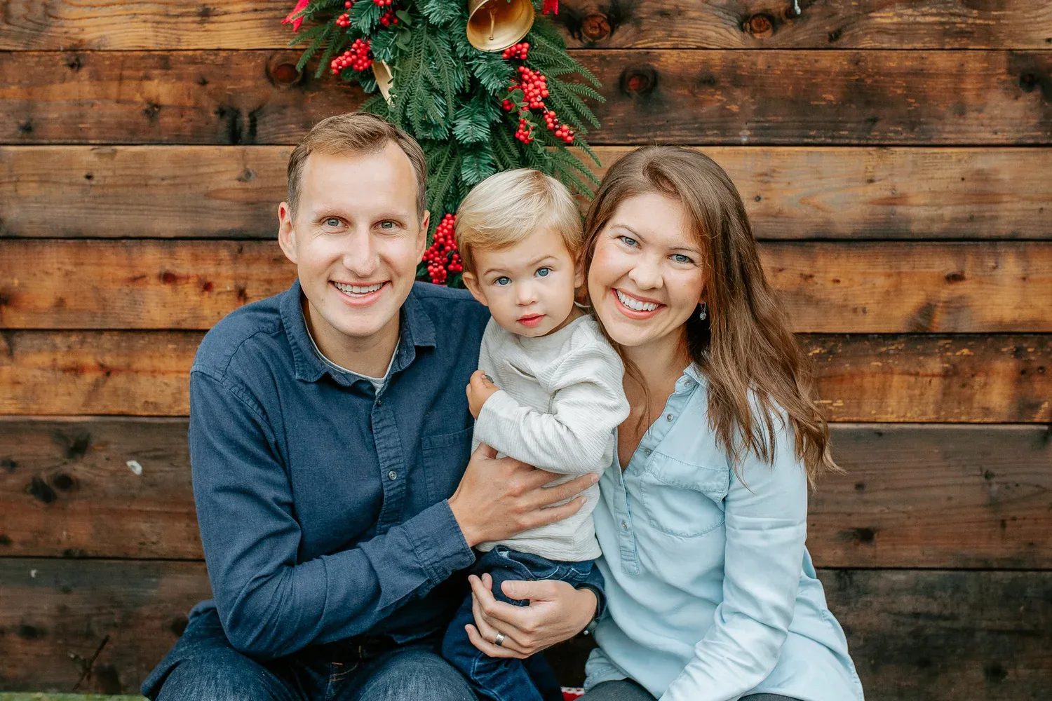 Two adults and a child sit in front of a wooden wall and holiday wreath