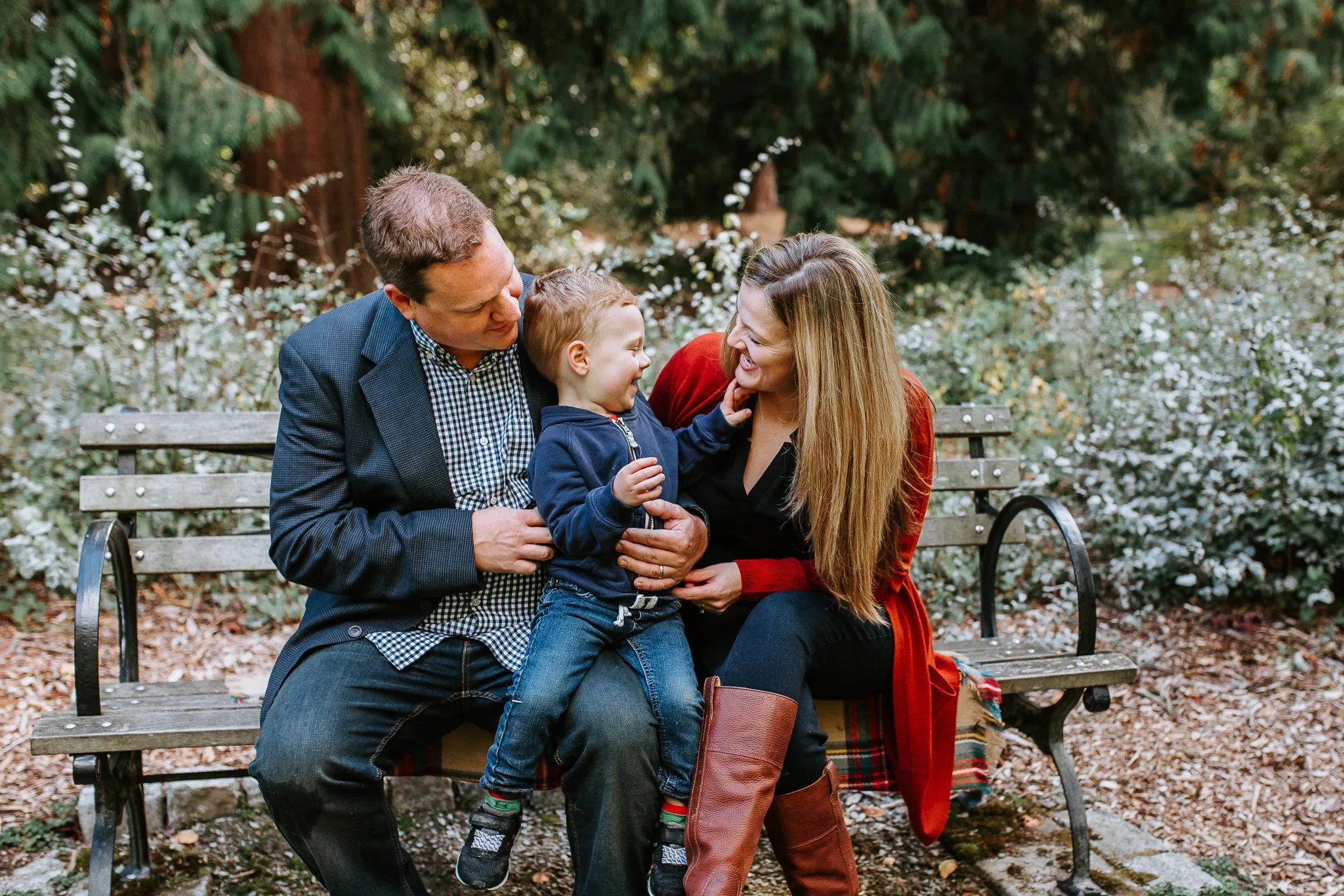 Two adults and a baby sit on a bench surrounded by autumn leaves