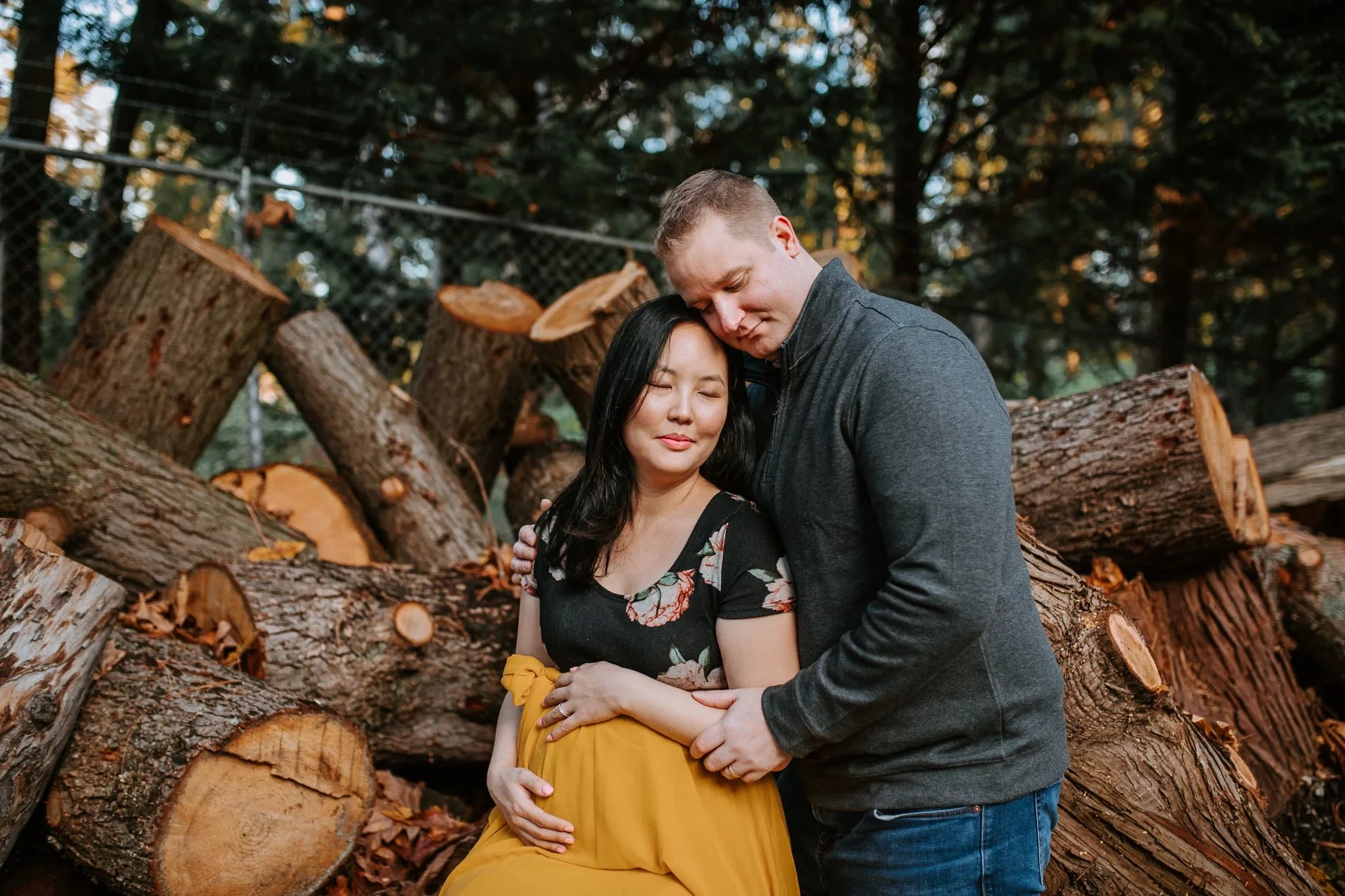 Two adults stand together beside a pile of cut logs in the woods