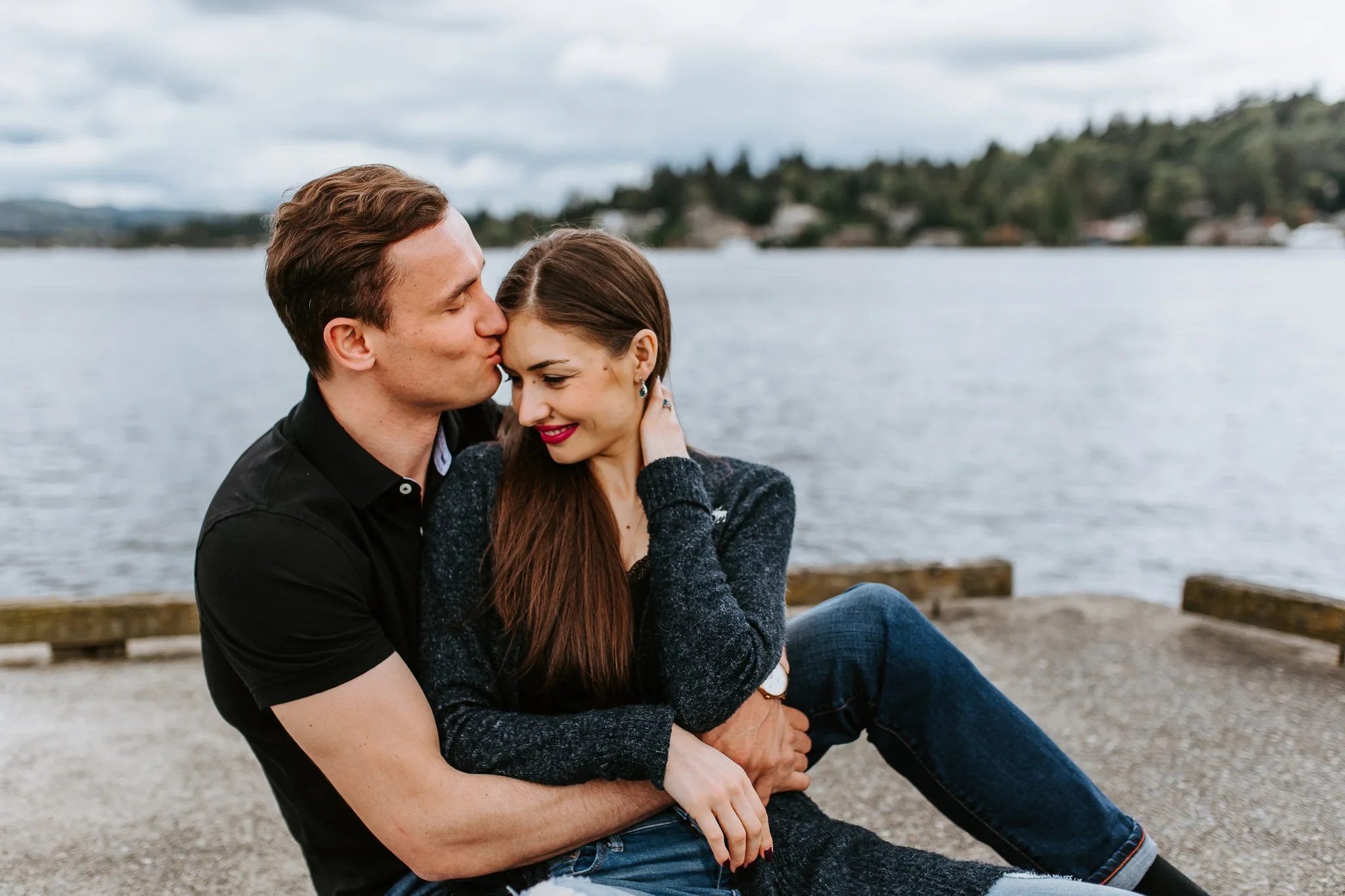 Two adults cuddle on a waterfront ledge with a black hat beside them
