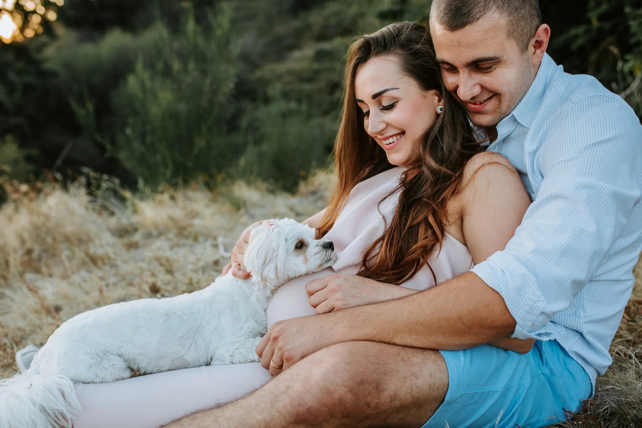 Two adults cuddle a white dog while sitting in dry grass