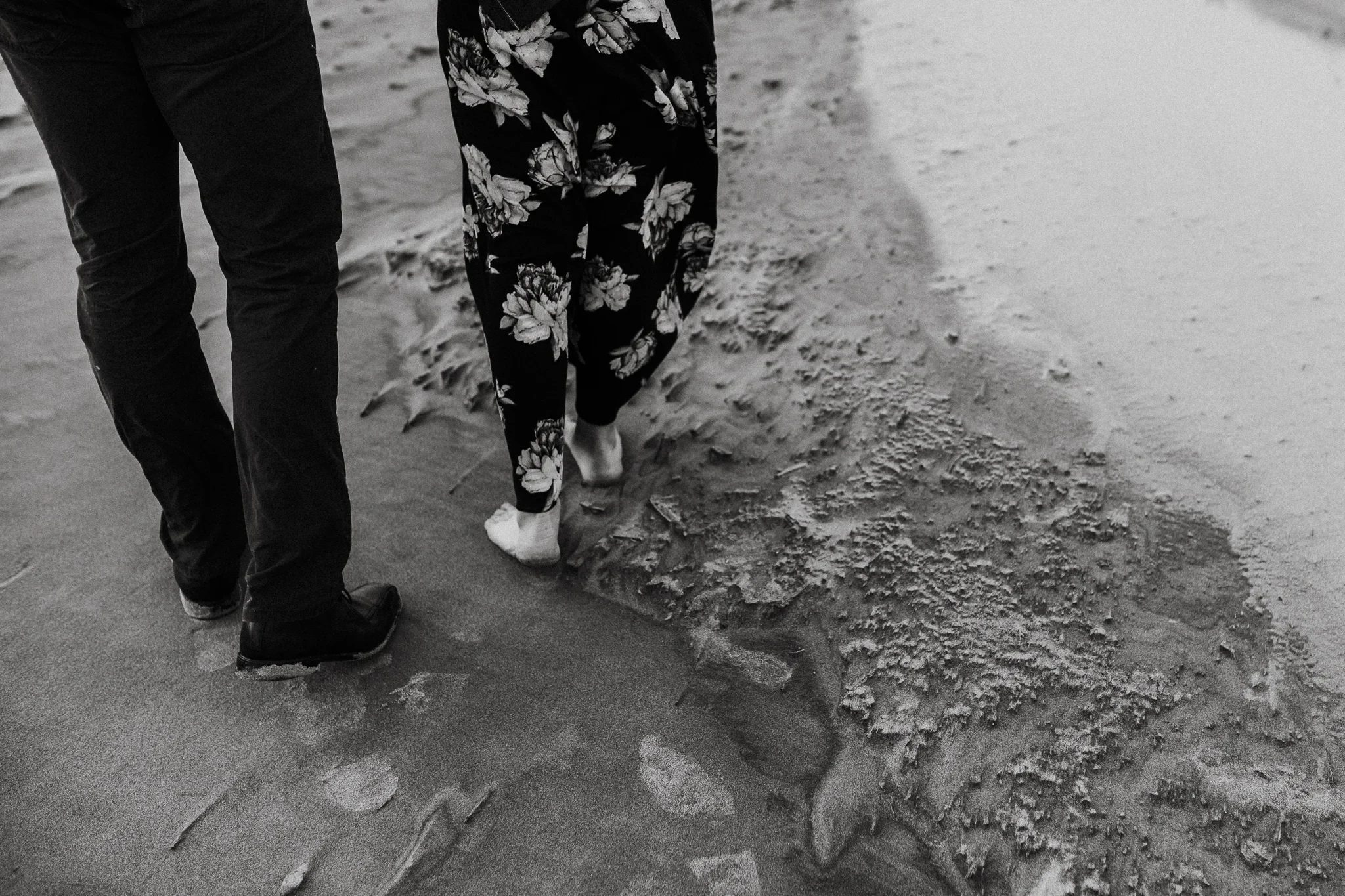 Black-and-white detail of two people walking barefoot along the wet shoreline