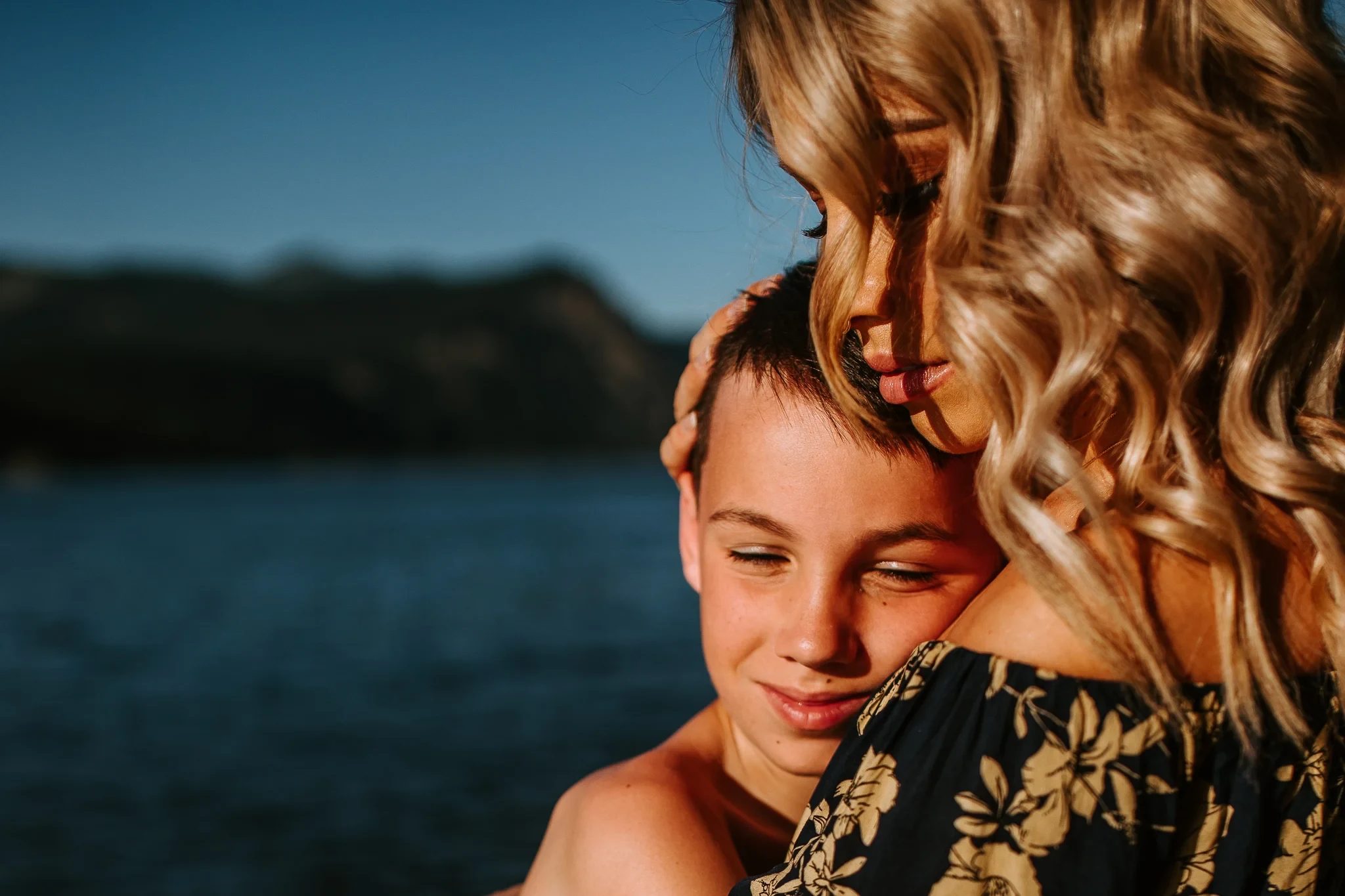 Child rests against a woman beside dark blue water