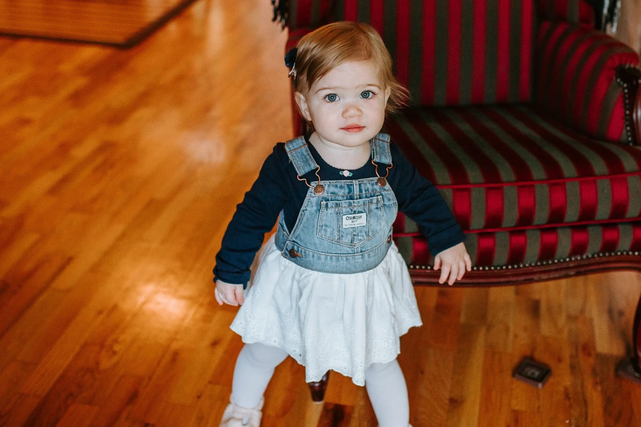 Child in a denim vest and pale dress stands on a wooden floor