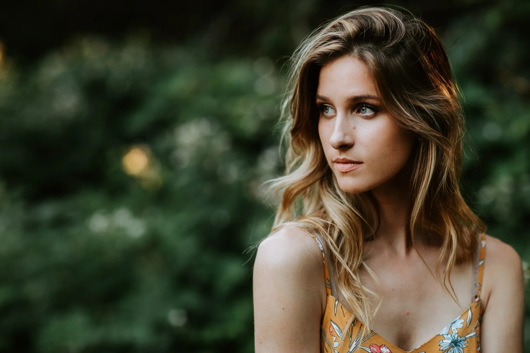 Woman in a floral top looks past the camera with soft green foliage behind her