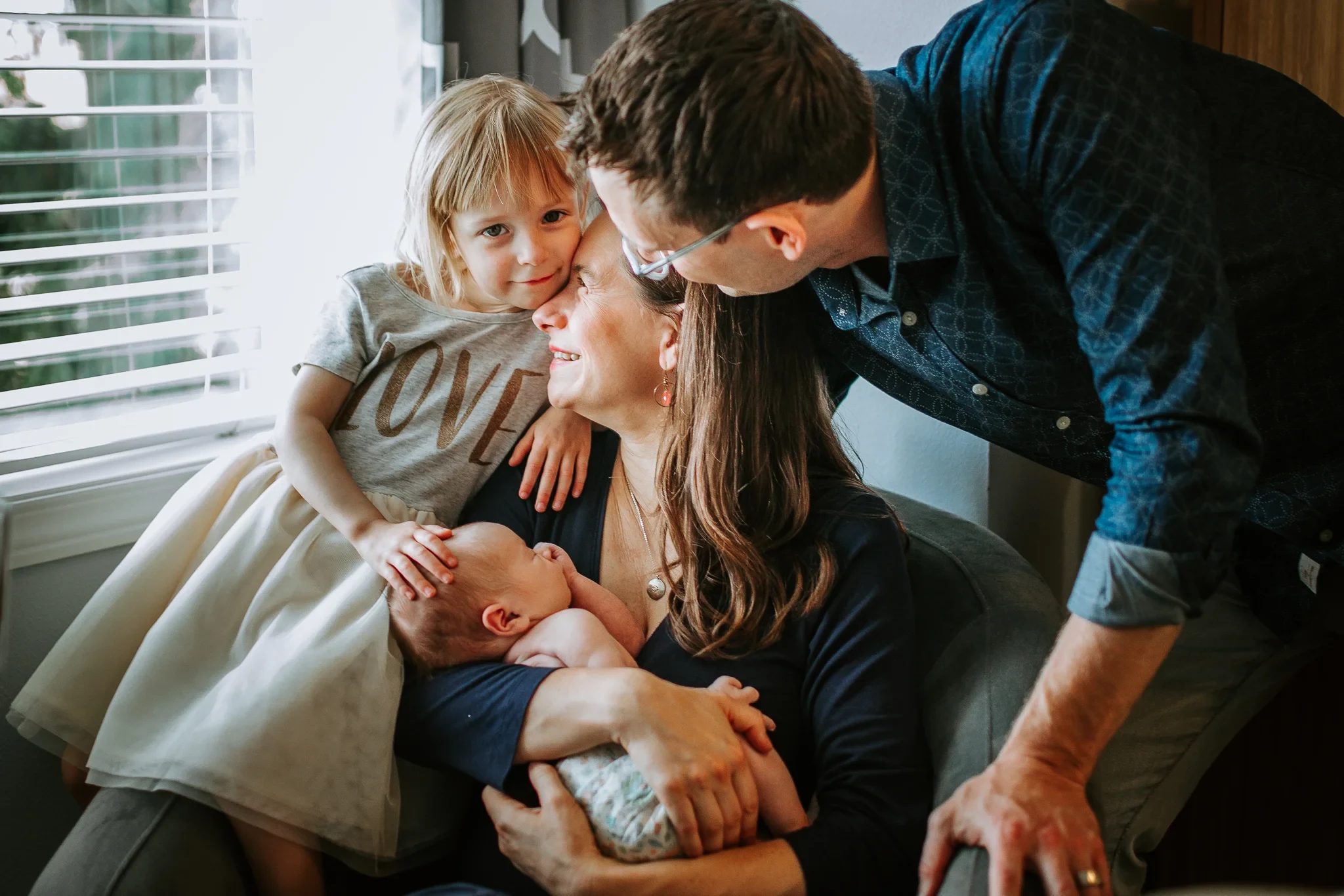 Two adults and a child gather around a baby beside a bright window
