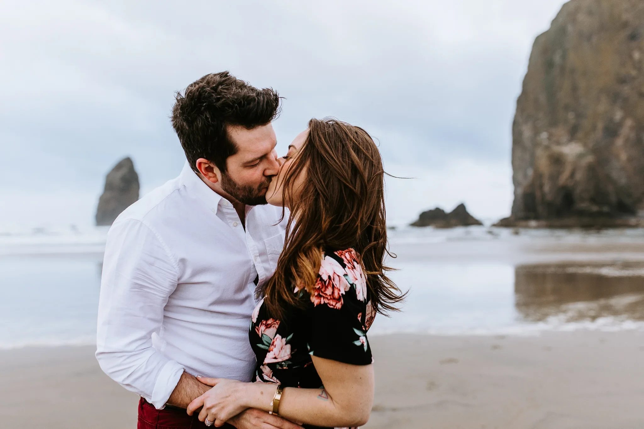 Two adults kiss on a sandy beach with a sea stack in the distance