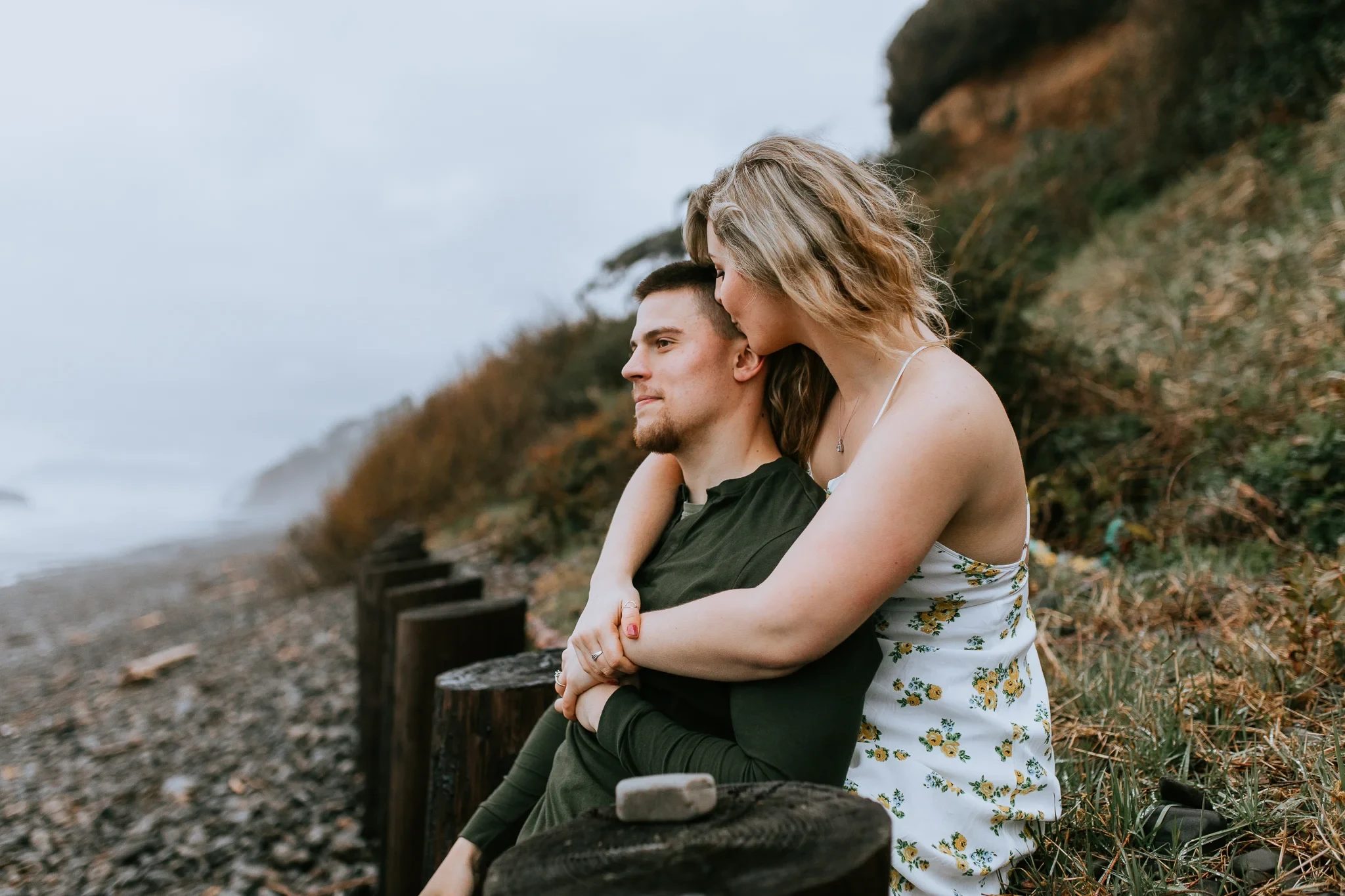Woman rests her arms around a man seated beside the shoreline