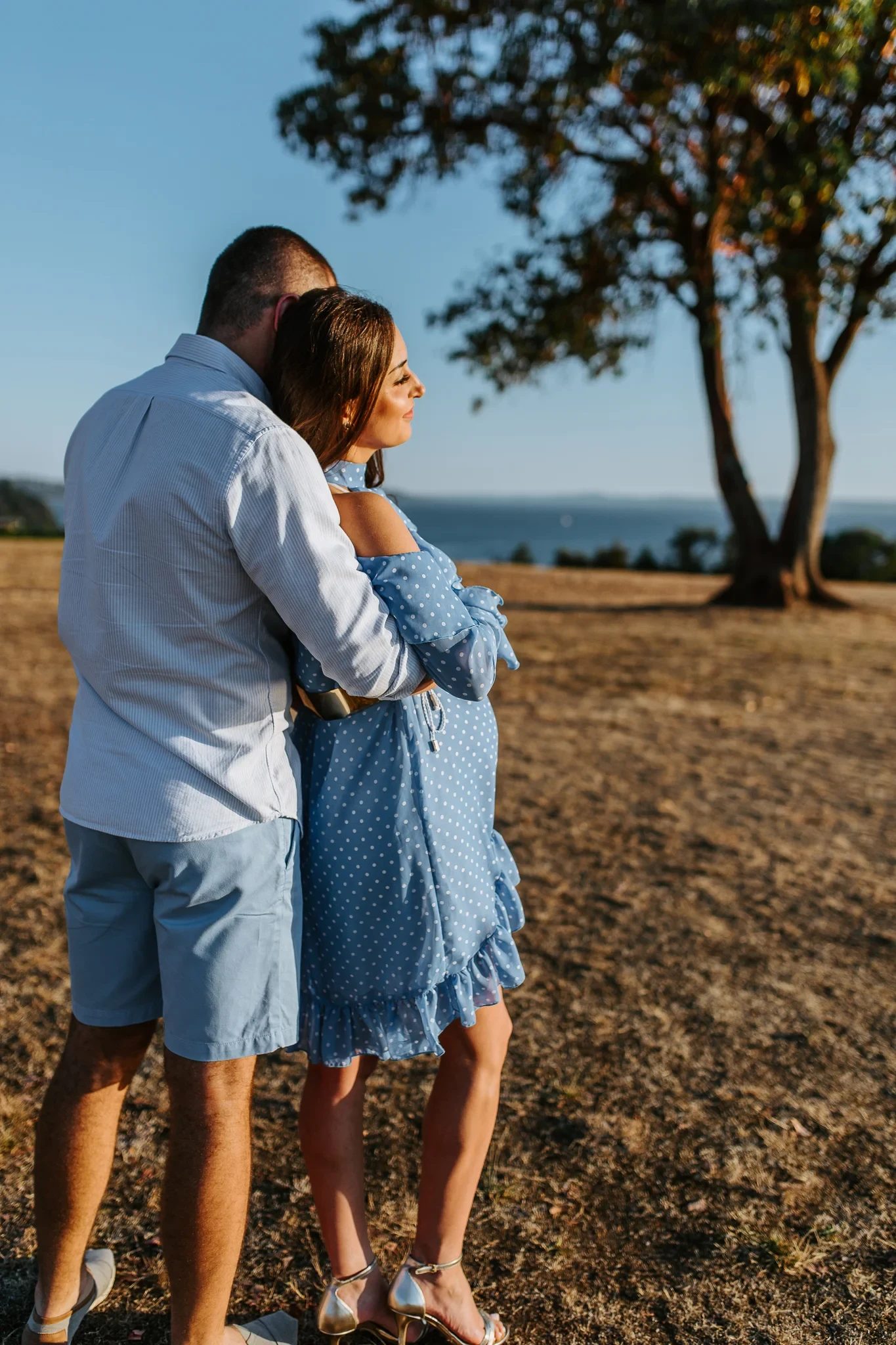 Two adults embrace beside a tree overlooking sunlit water