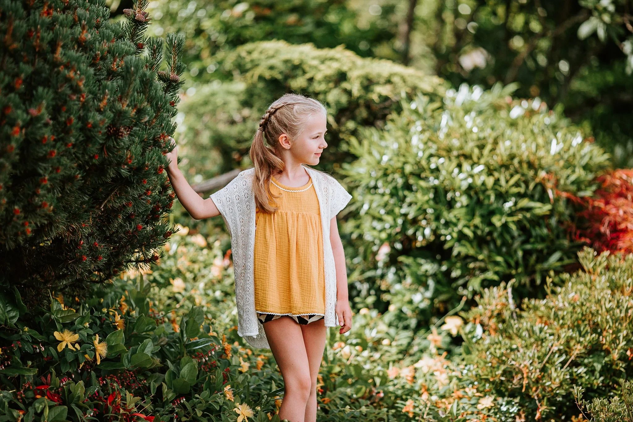 Child in a yellow top stands beside dense green garden shrubs