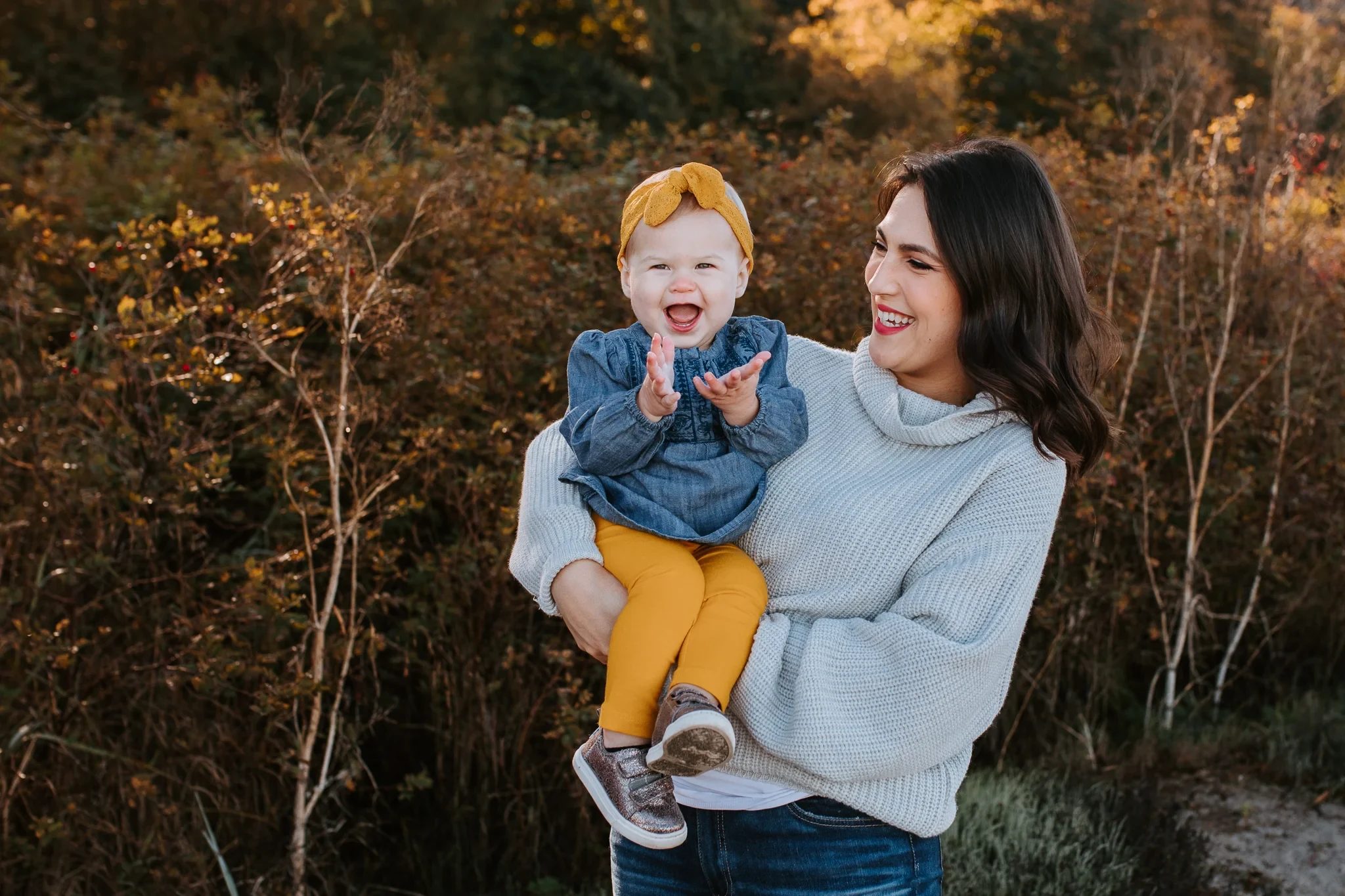 Woman holds a child wearing a yellow bow among autumn shrubs