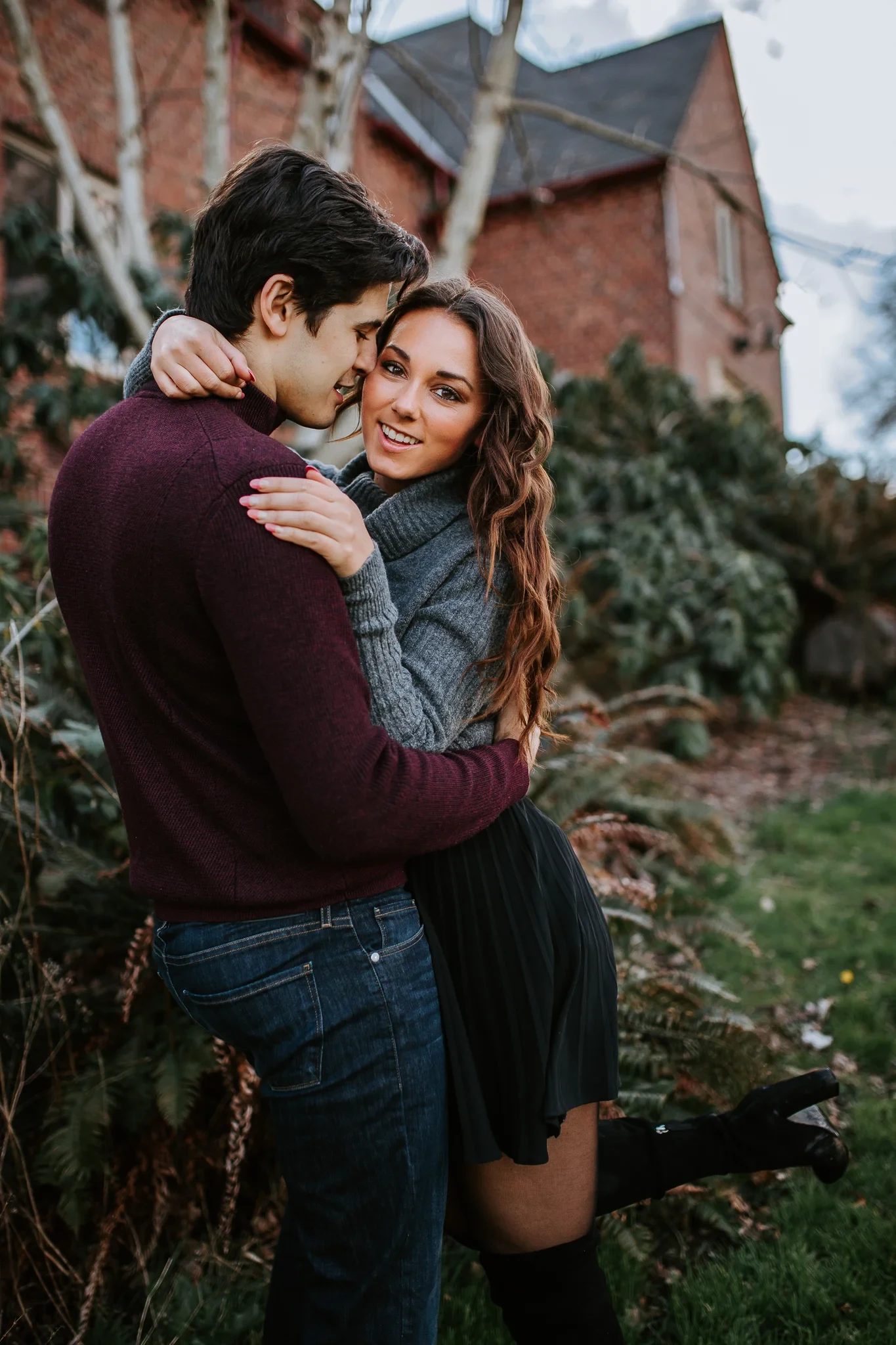 Two adults embrace beside a brick building and autumn plants