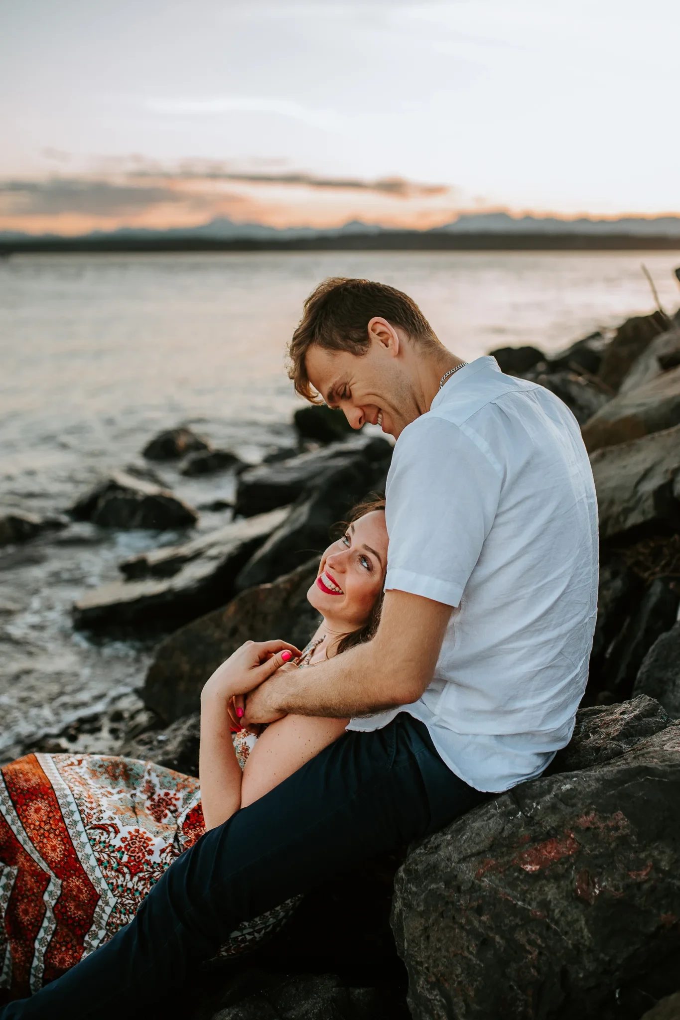 Woman leans back against a man while seated on shoreline rocks
