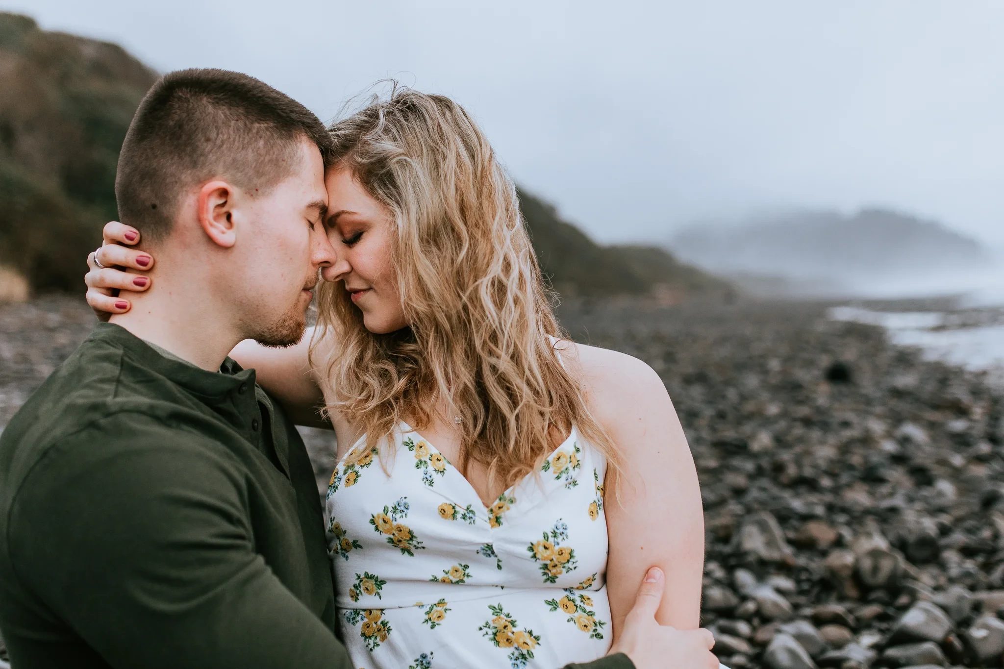 Two adults touch foreheads on a rocky beach beneath a cloudy sky