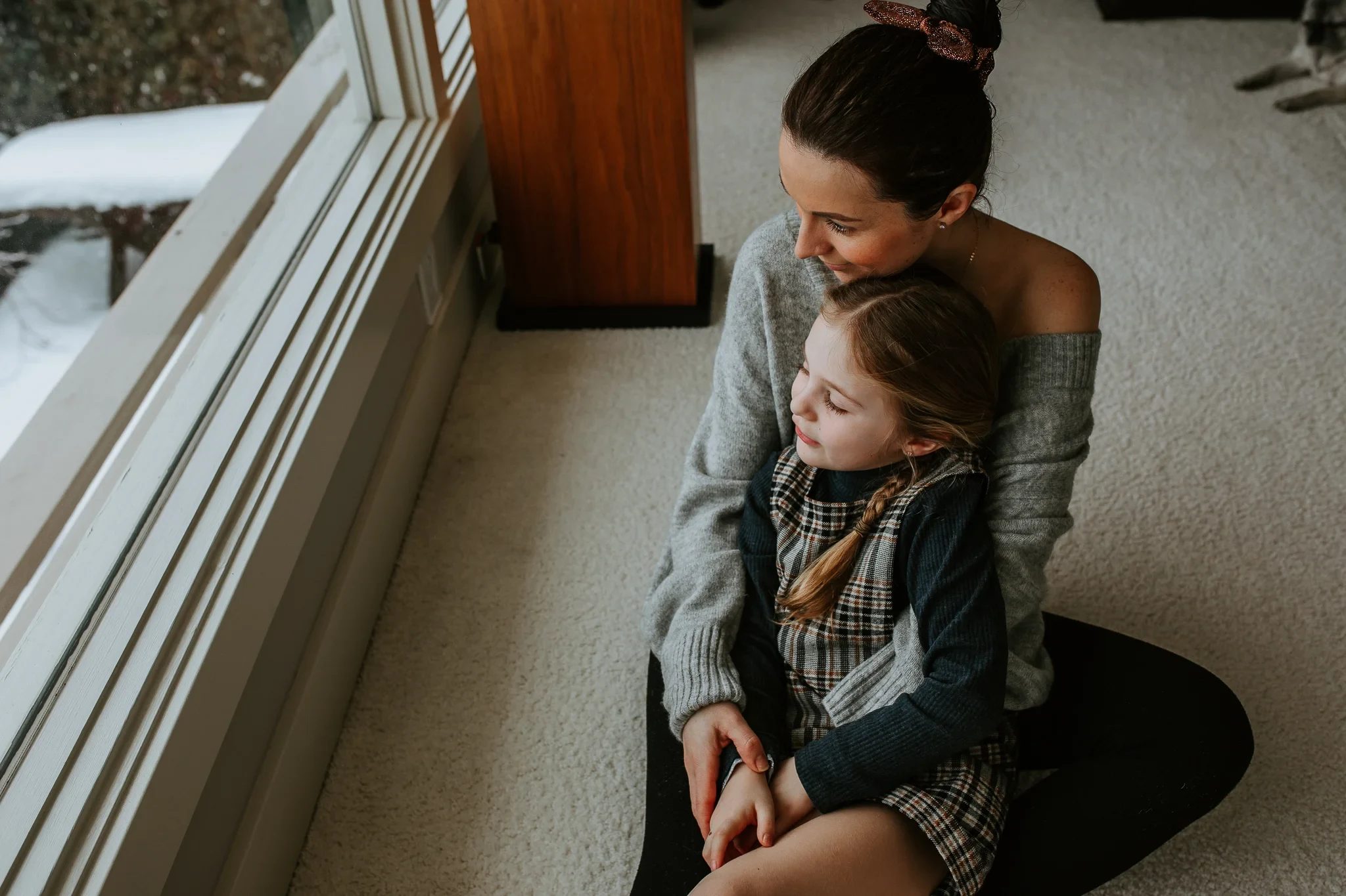 Woman sits on the floor holding a child close beside a window