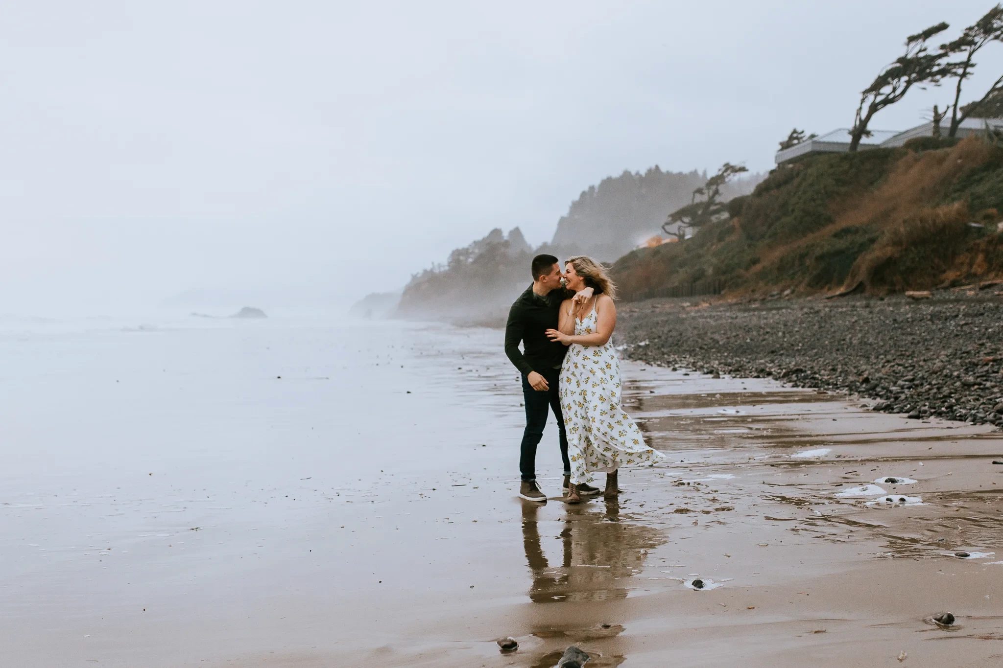 Two adults embrace at the water’s edge beneath a misty wooded bluff