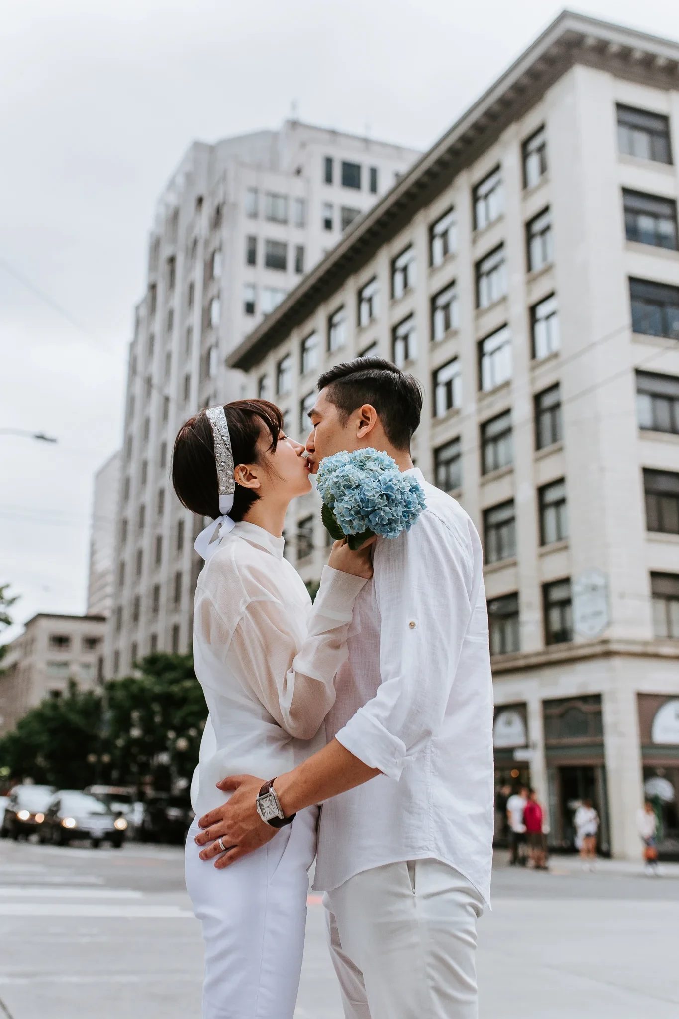 Two adults in white clothing embrace beneath tall city buildings