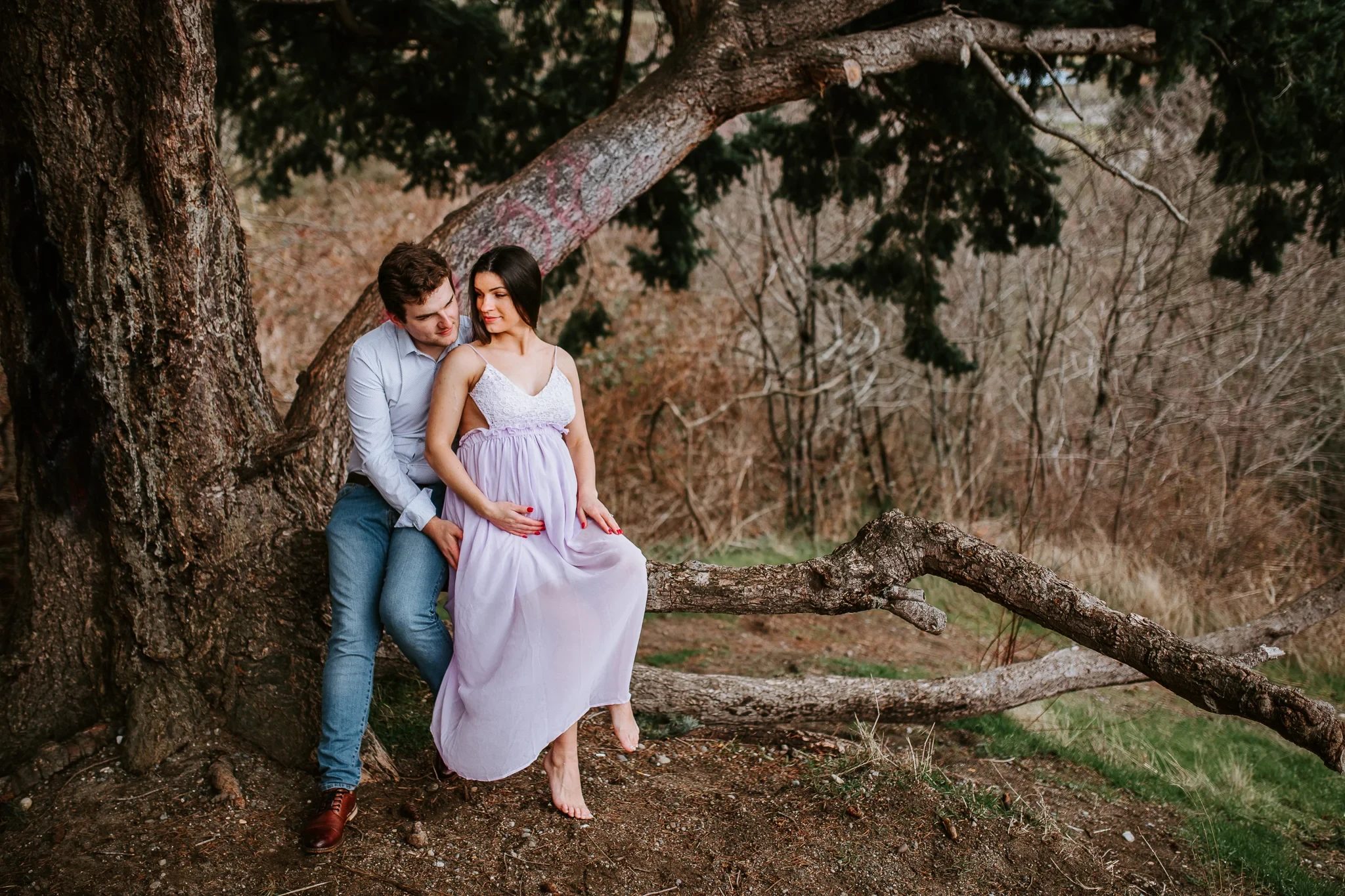 Two adults sit beneath a wide tree beside a fallen branch