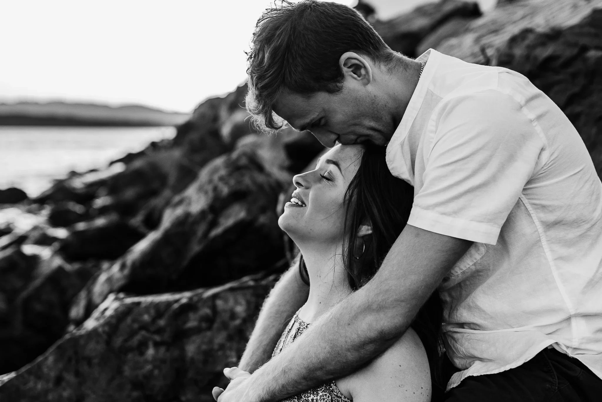 Black-and-white portrait of two adults leaning together on shoreline rocks