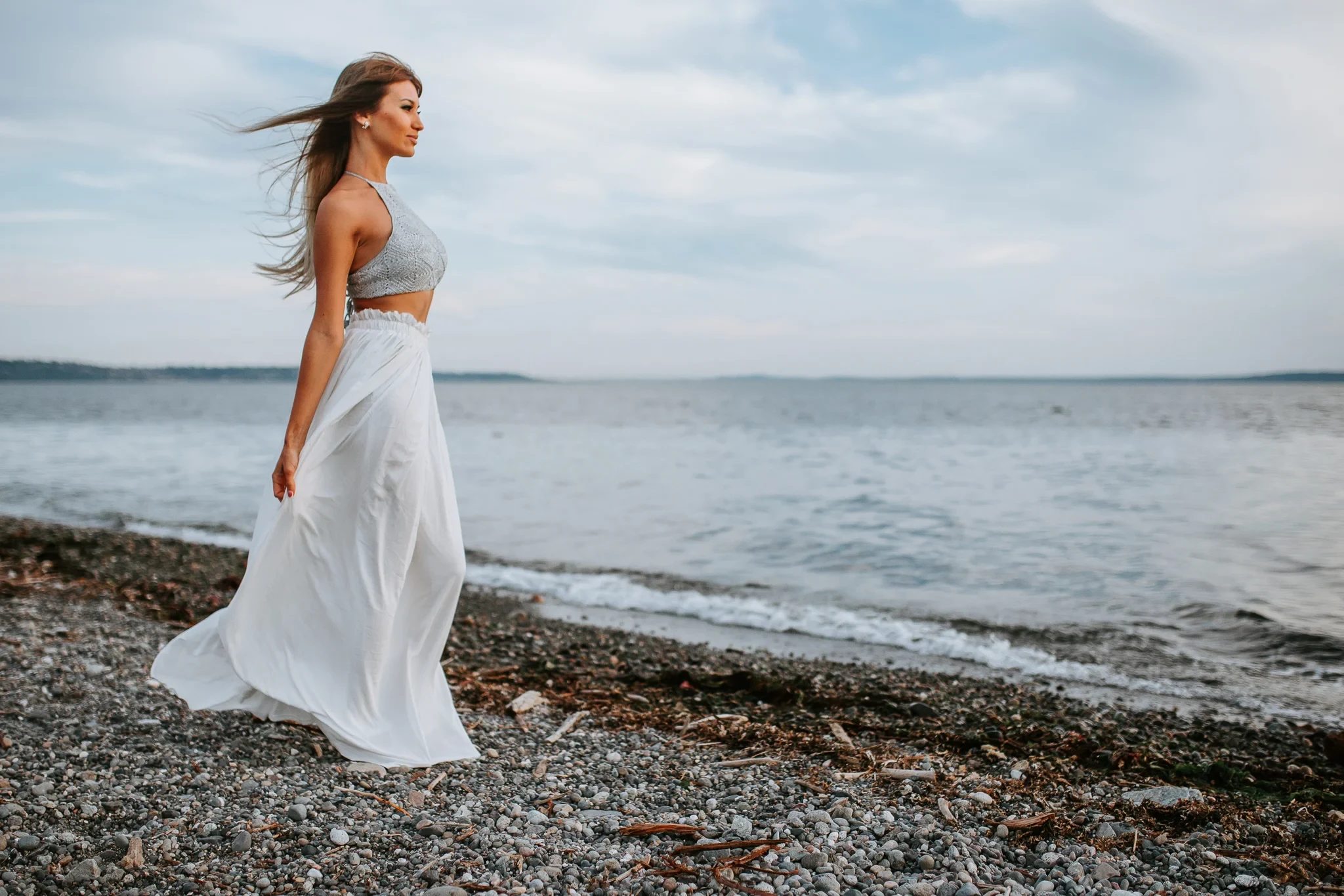 Woman in a white dress turns toward the water on a pebbled shore
