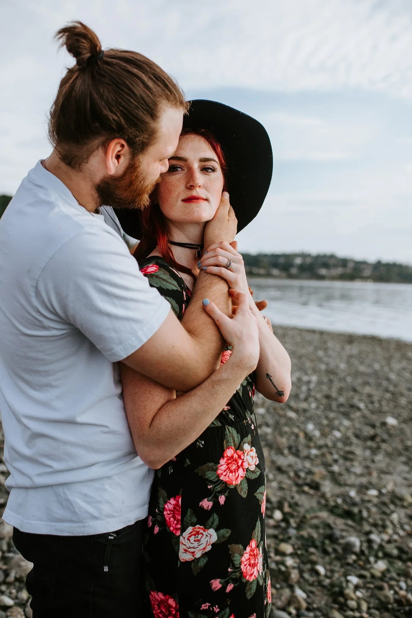 Two adults embrace on the beach, one wearing a black hat