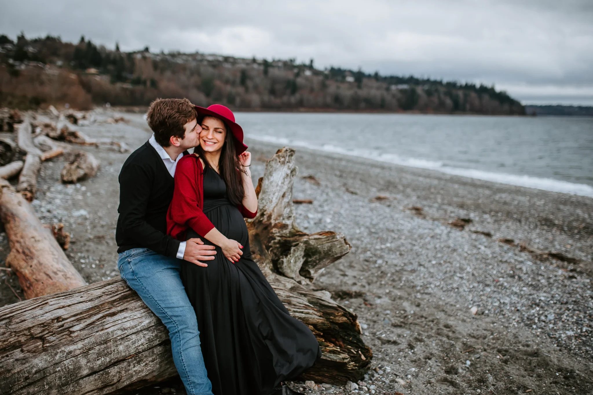 Two adults sit together on driftwood beside a broad stretch of shoreline