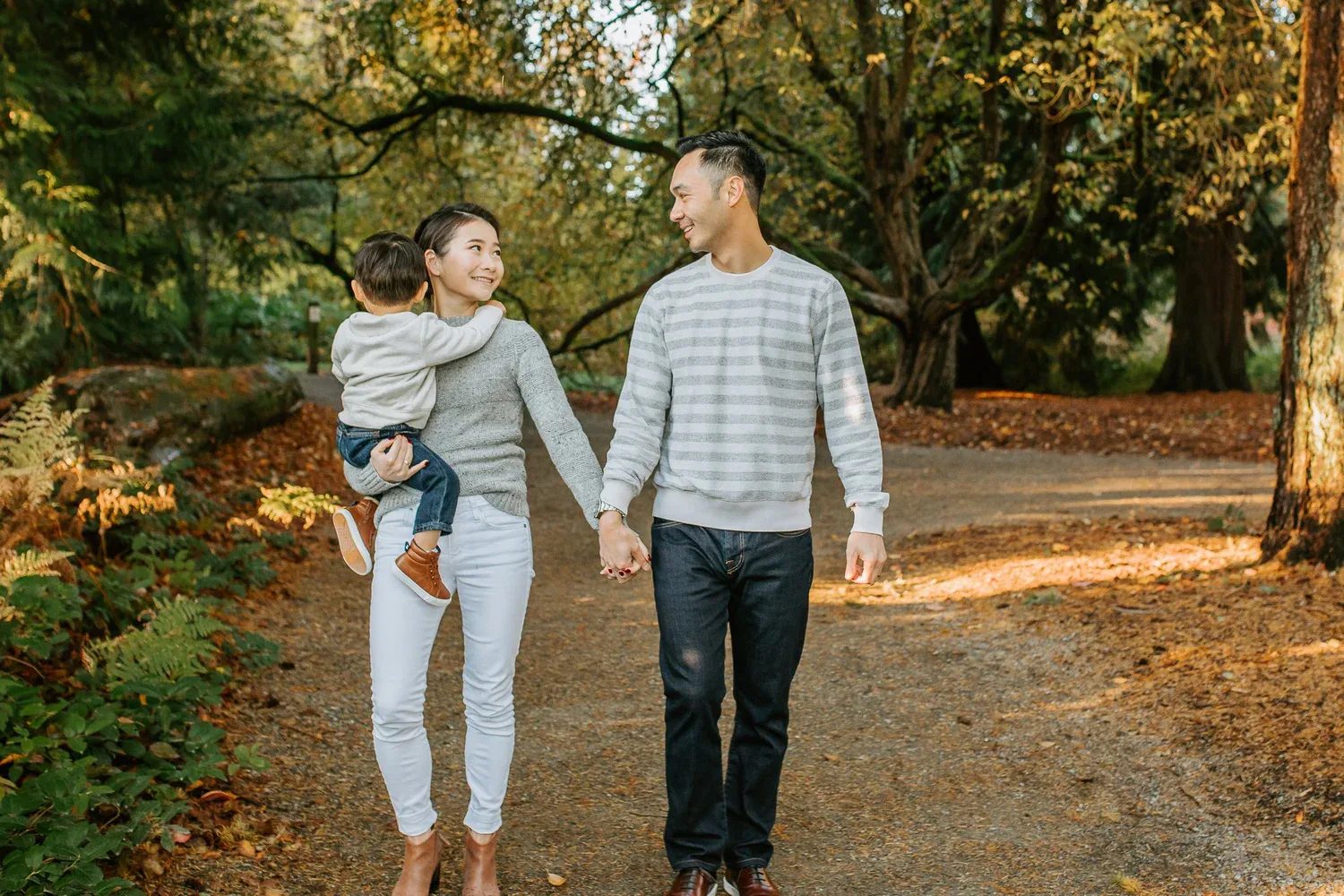 Two adults walk hand in hand while carrying a child along a leafy path
