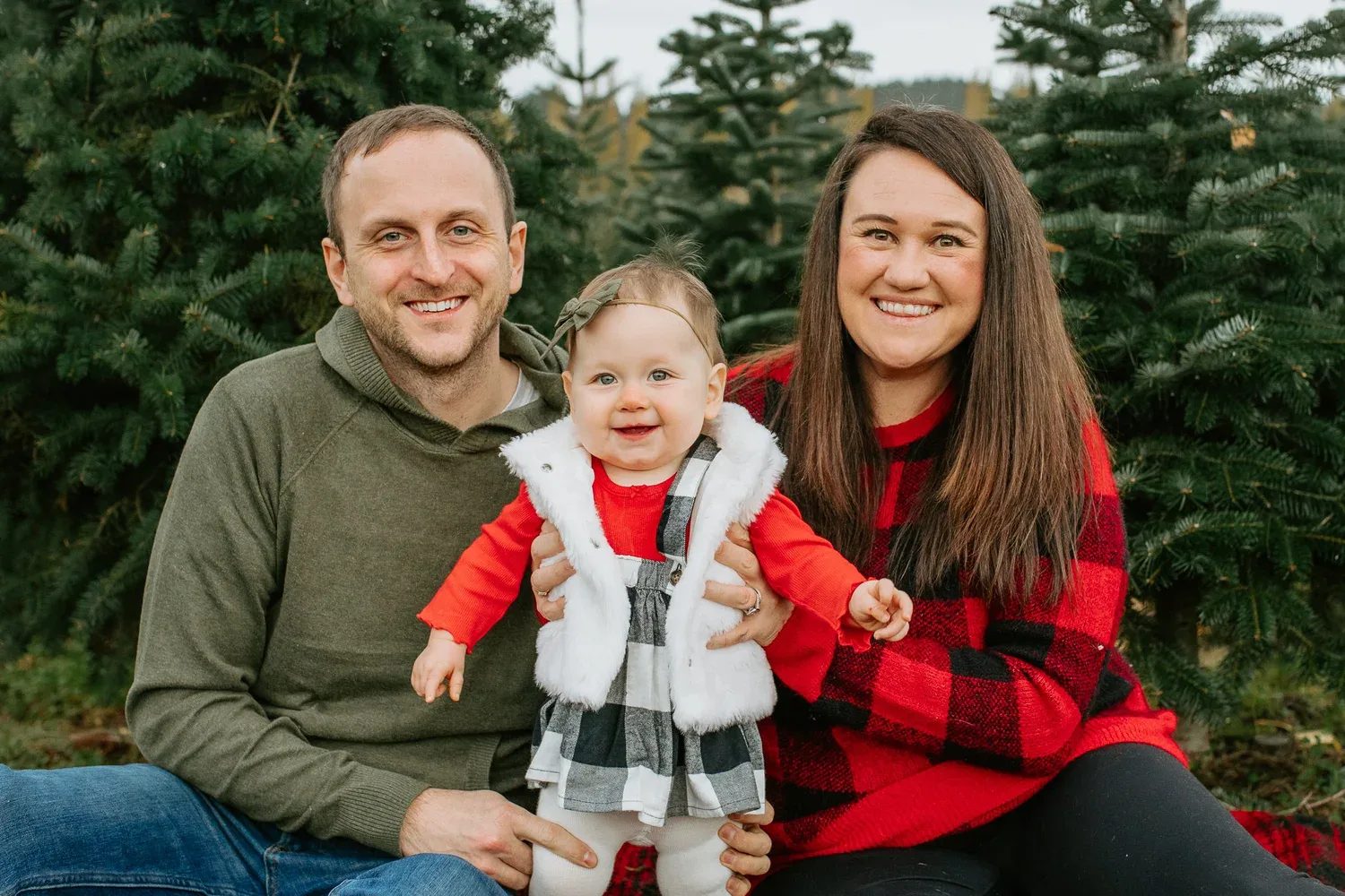 Two adults sit with a child in red clothing among evergreen trees