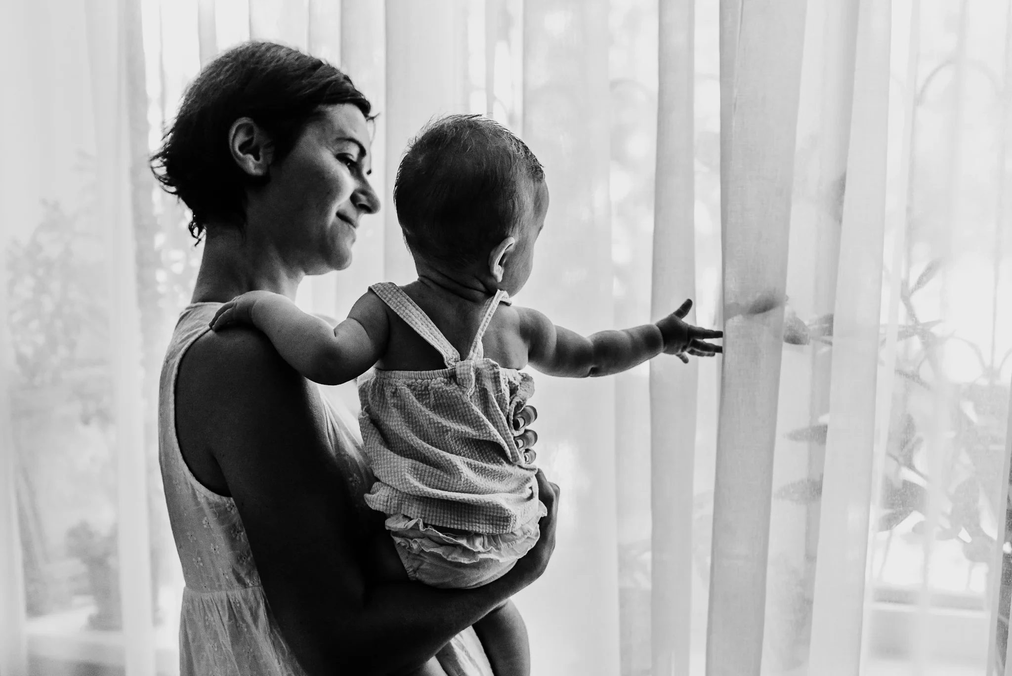 Black-and-white silhouette of a woman holding a child beside sheer curtains