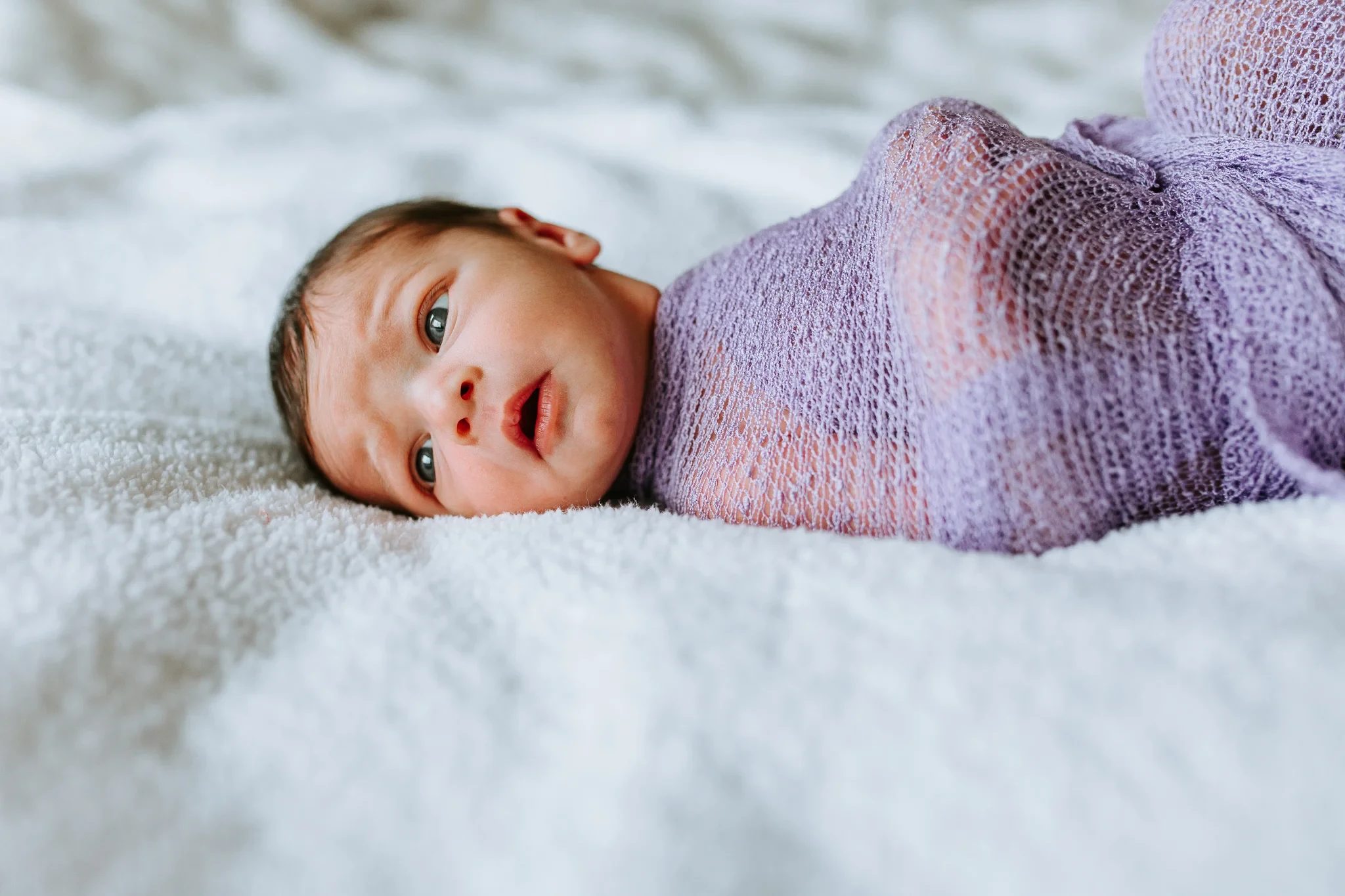 Baby wrapped in a lavender blanket looks toward the camera