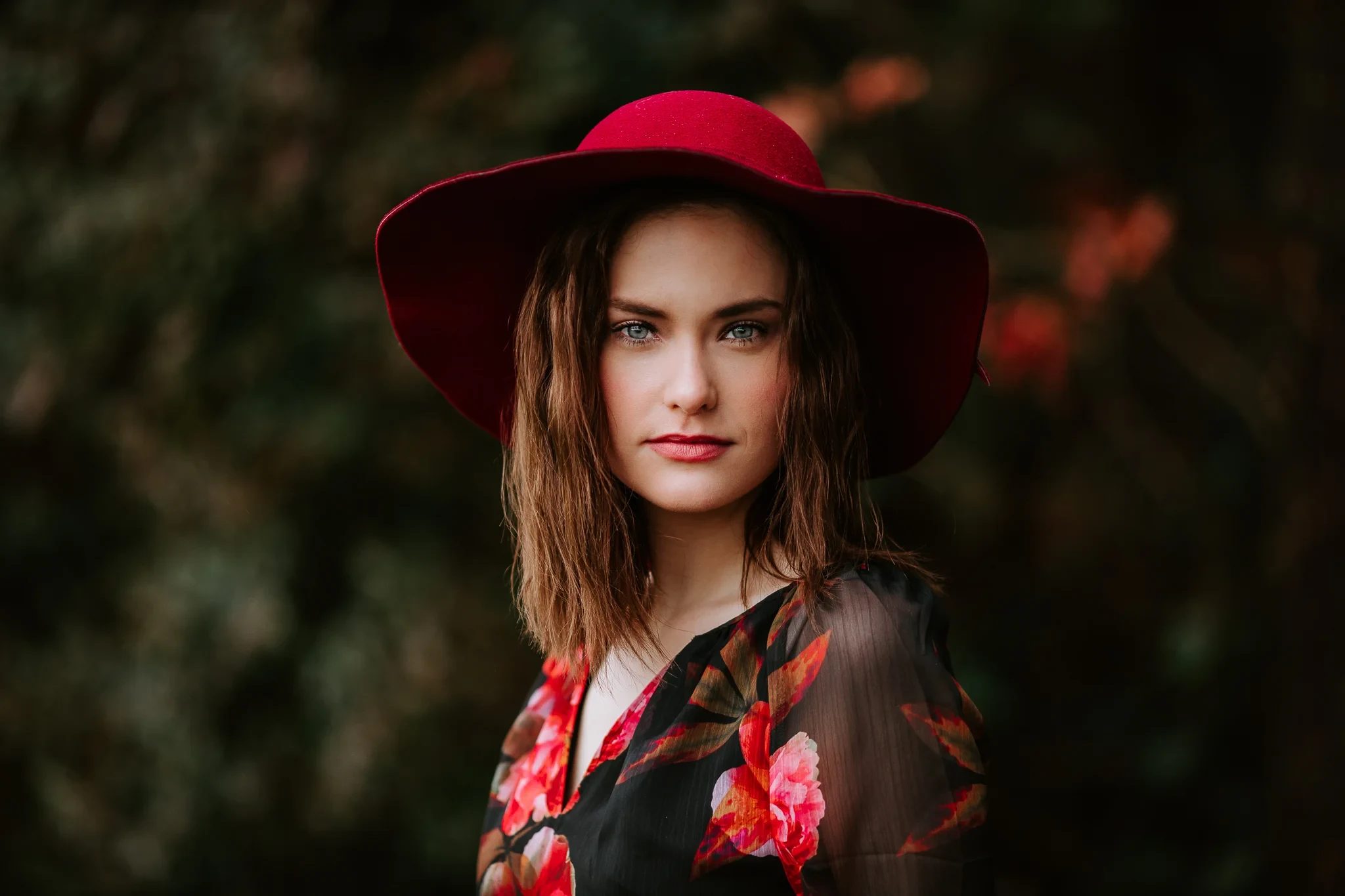 Woman wearing a wide red hat and floral dress looks toward the camera