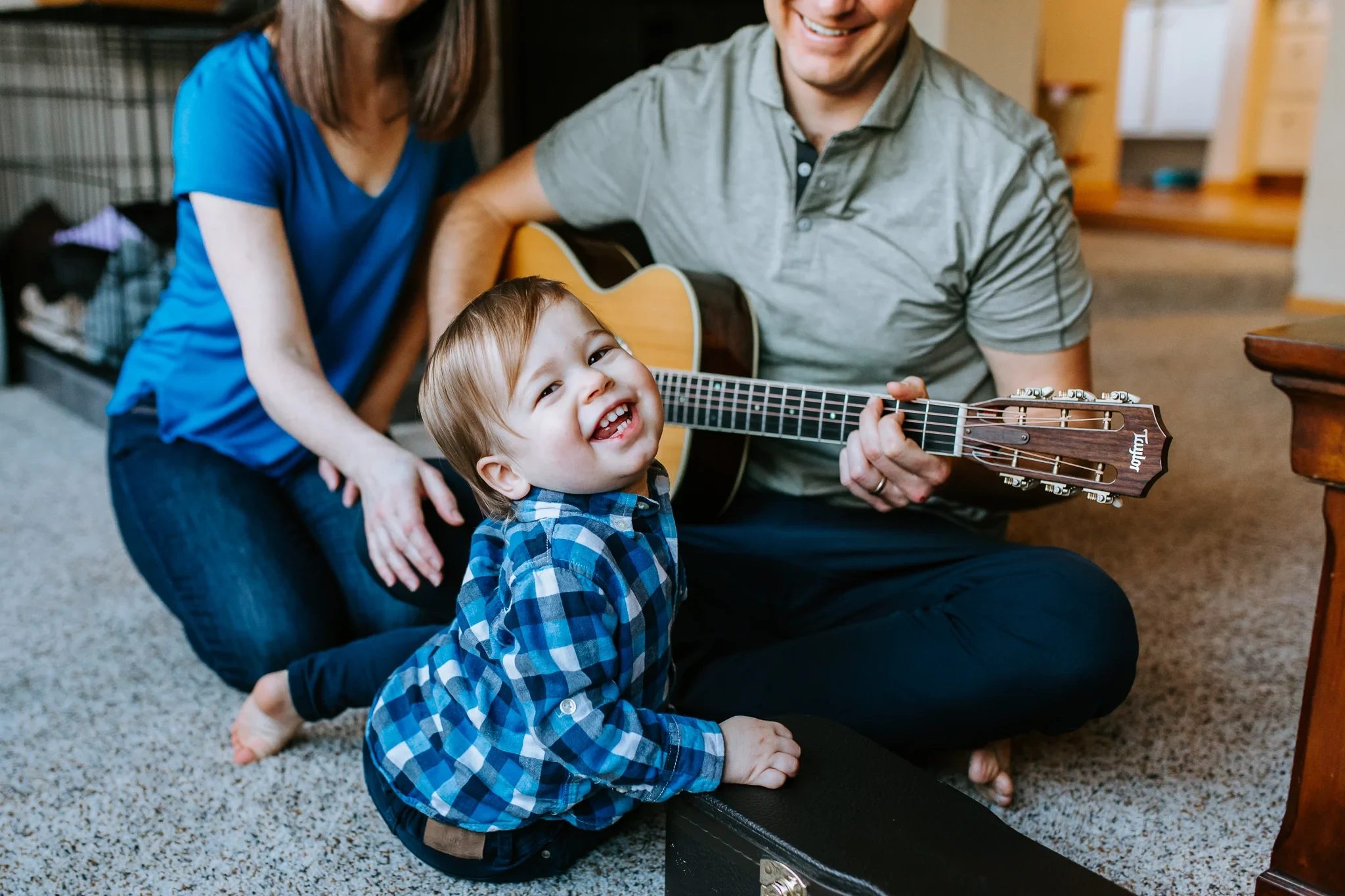 Child laughs beside two adults playing an acoustic guitar