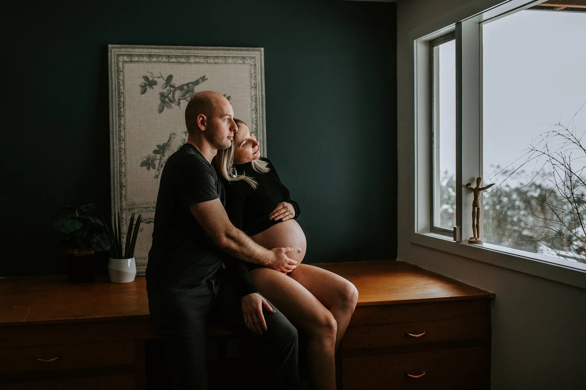 Two adults sit together on a wooden ledge beside a large window