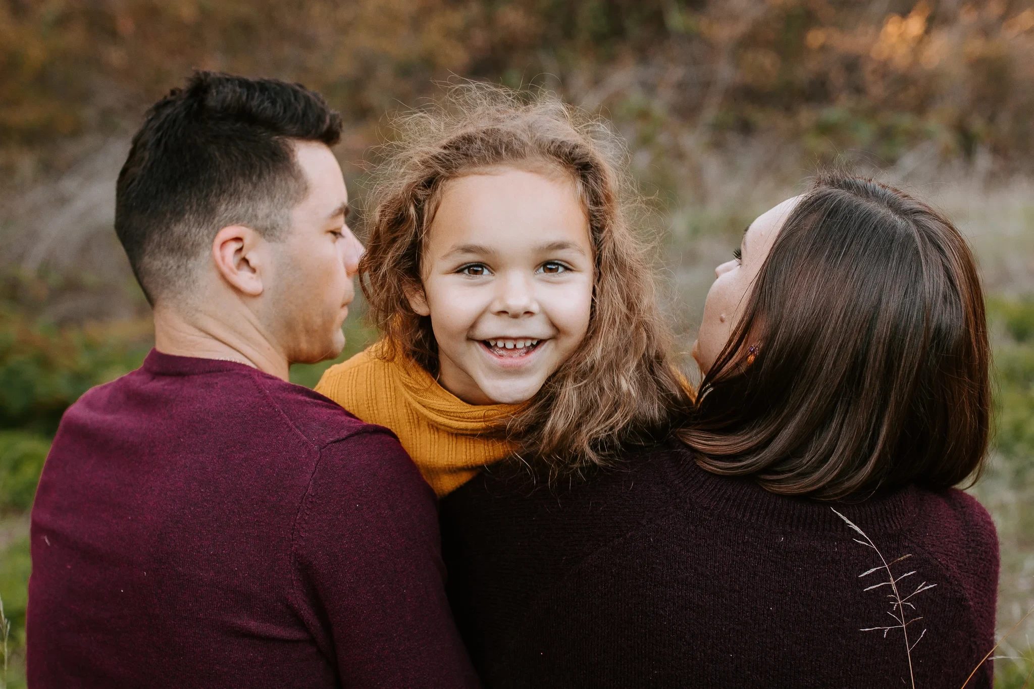 Child smiles between two adults with autumn trees behind them