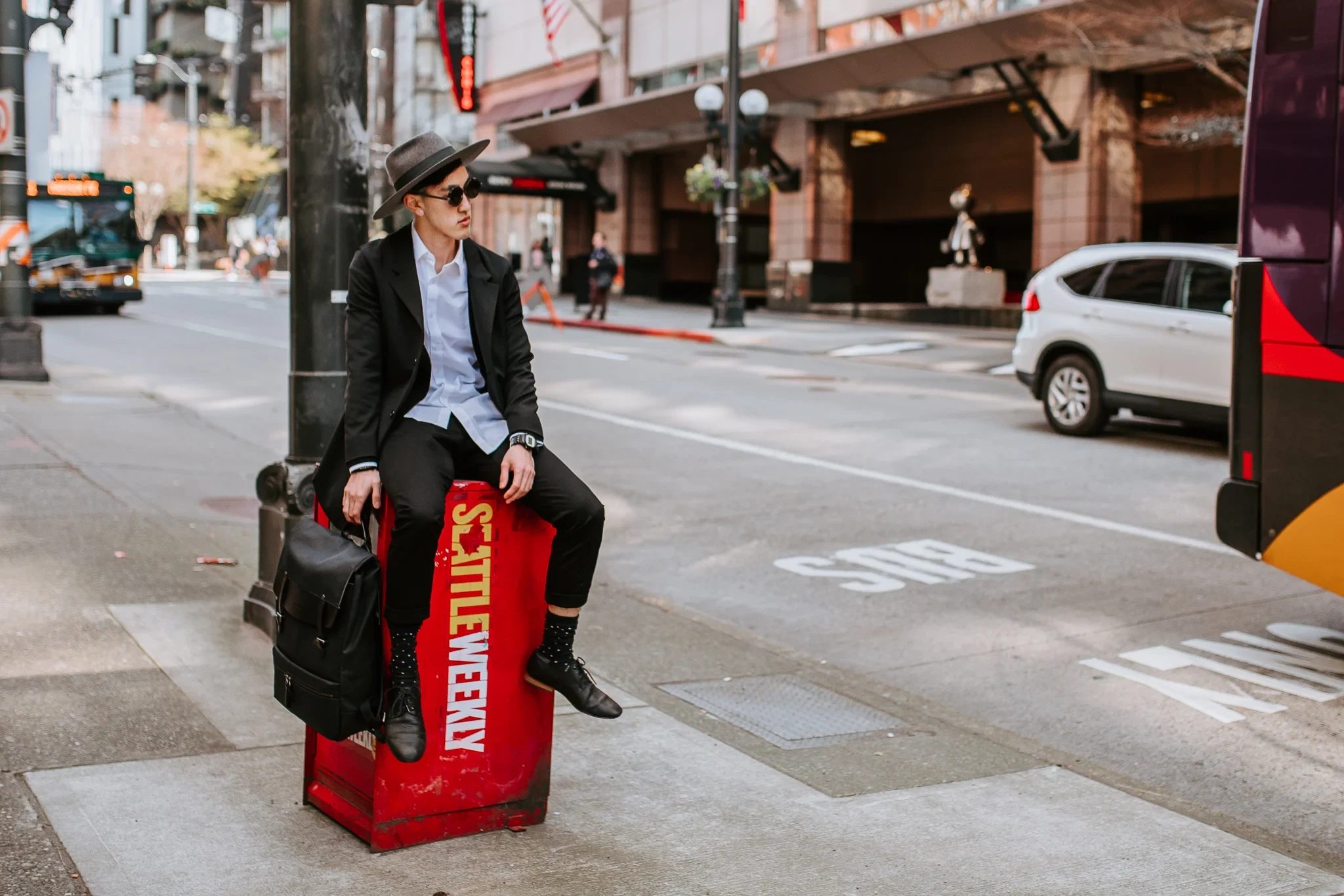 Person in a hat sits on a red newspaper box beside a city street