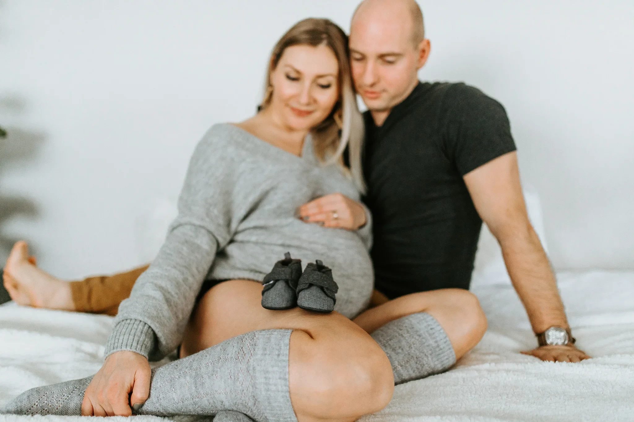 Two adults sit on a bed with small shoes resting on a grey dress