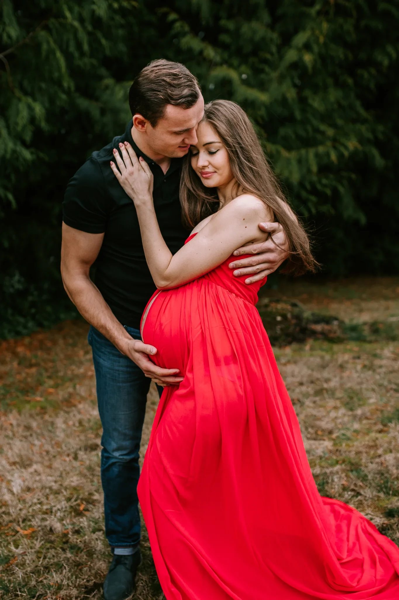 Two adults embrace beneath trees, with a long red dress in the foreground